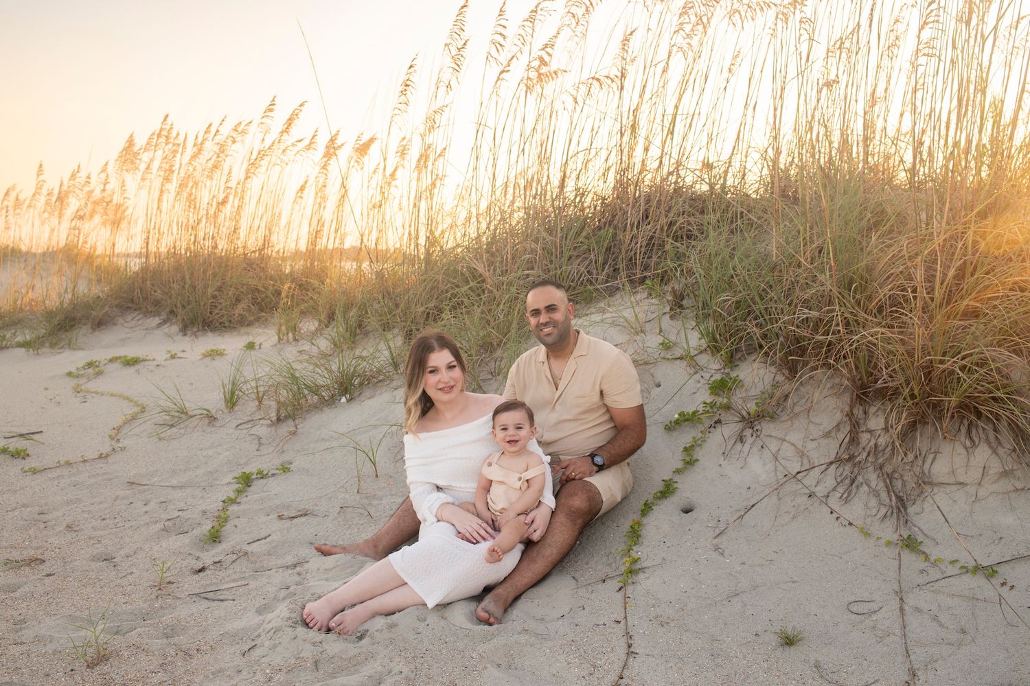 Family of three sitting on sandy beach dunes at golden sunset, parents smiling with baby in neutral tones.