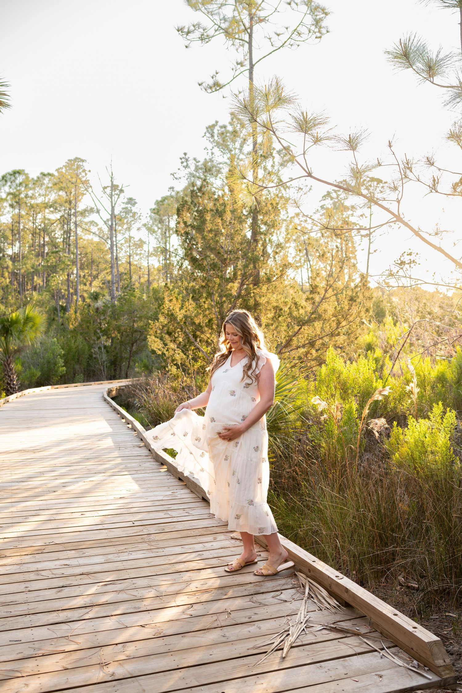Pregnant woman in white floral dress standing on wooden boardwalk surrounded by lush greenery in golden sunlight.