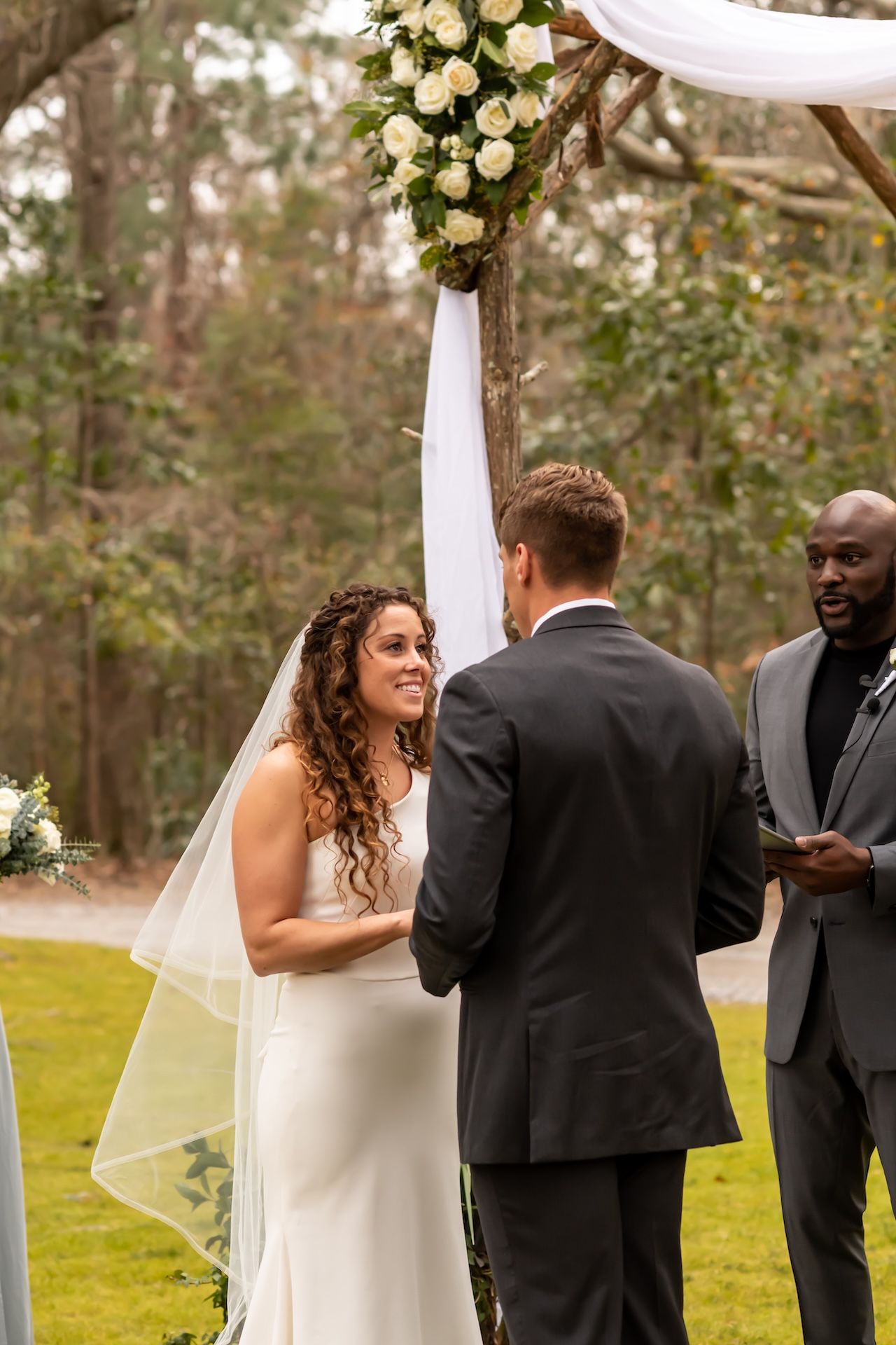 Bride and groom exchanging vows outdoors under a floral arch, officiant holding book, forest backdrop.