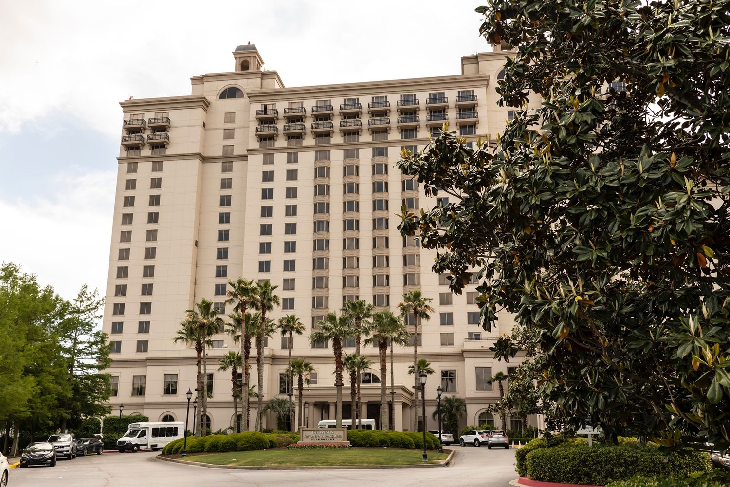 A tall luxury hotel building with white facade and arched windows stands surrounded by palm trees and landscaping.