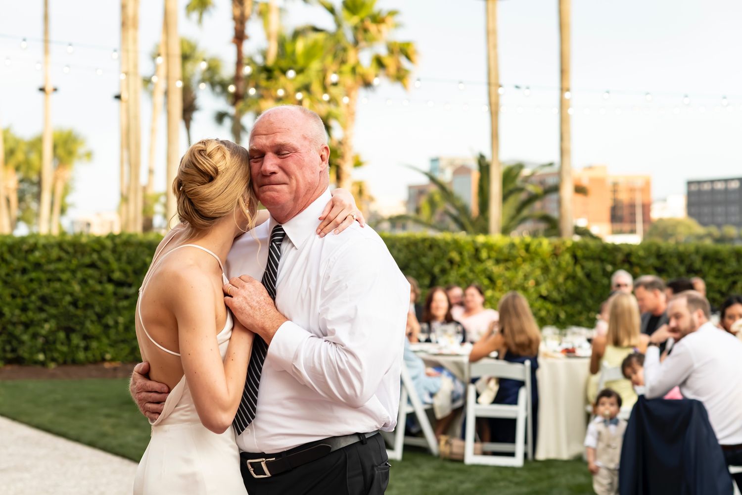 A tender dance moment captured at an outdoor wedding reception with palm trees and white chairs in the background.