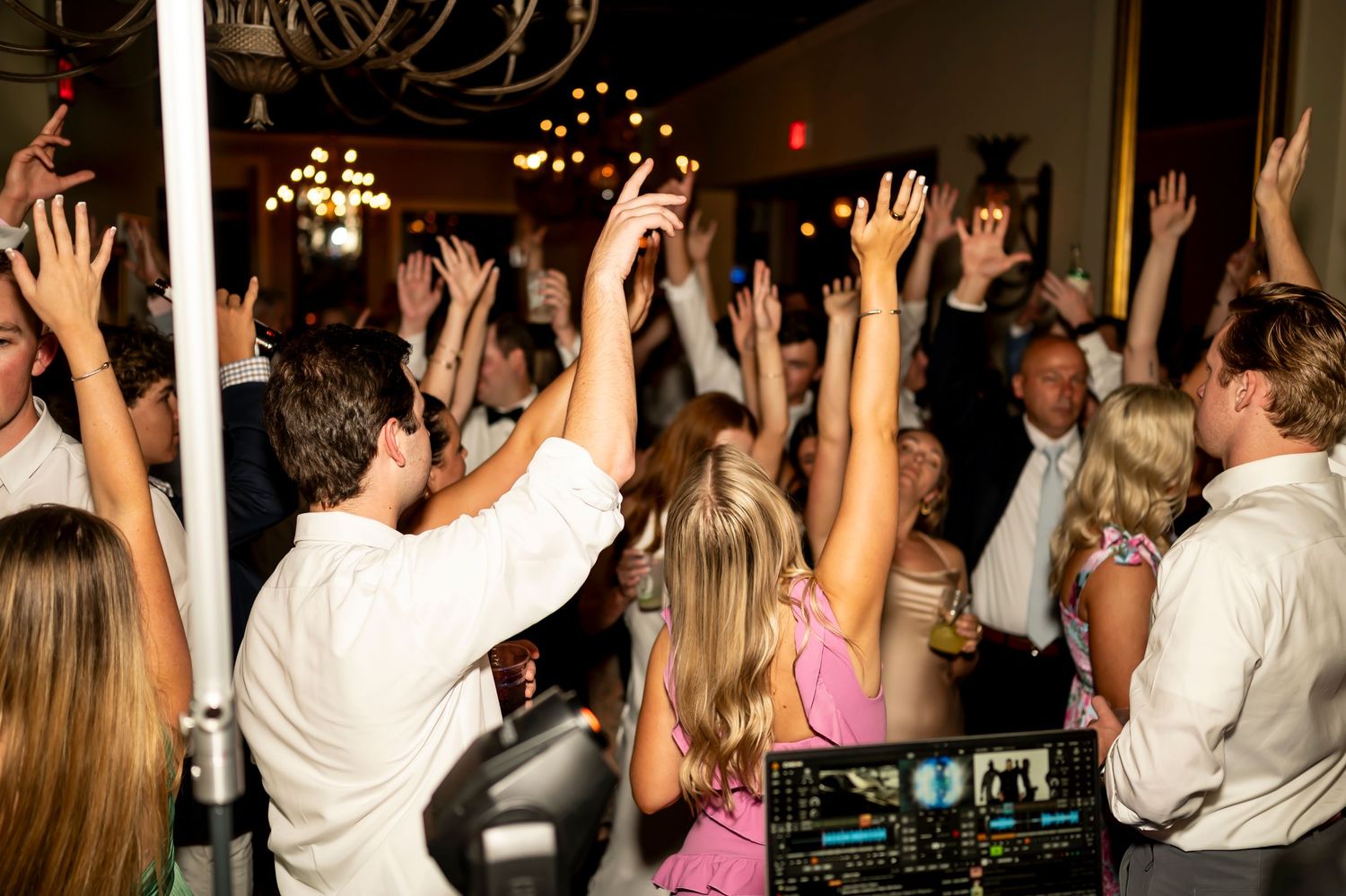 Crowded nightclub dance floor with people raising their hands in celebration as DJ performs at booth.