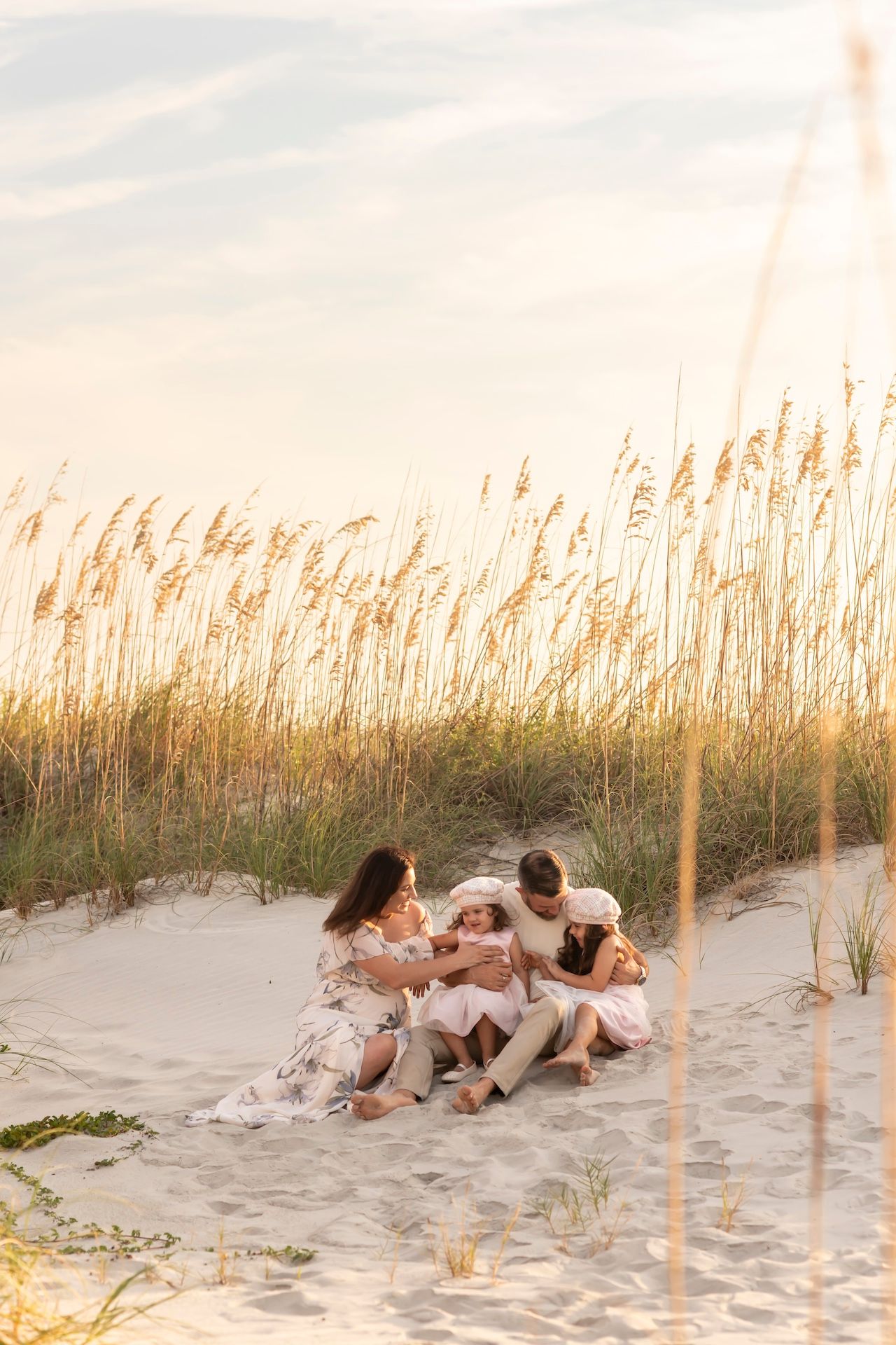 A family sits together on a sandy beach at sunset with sea oats swaying in the golden light.