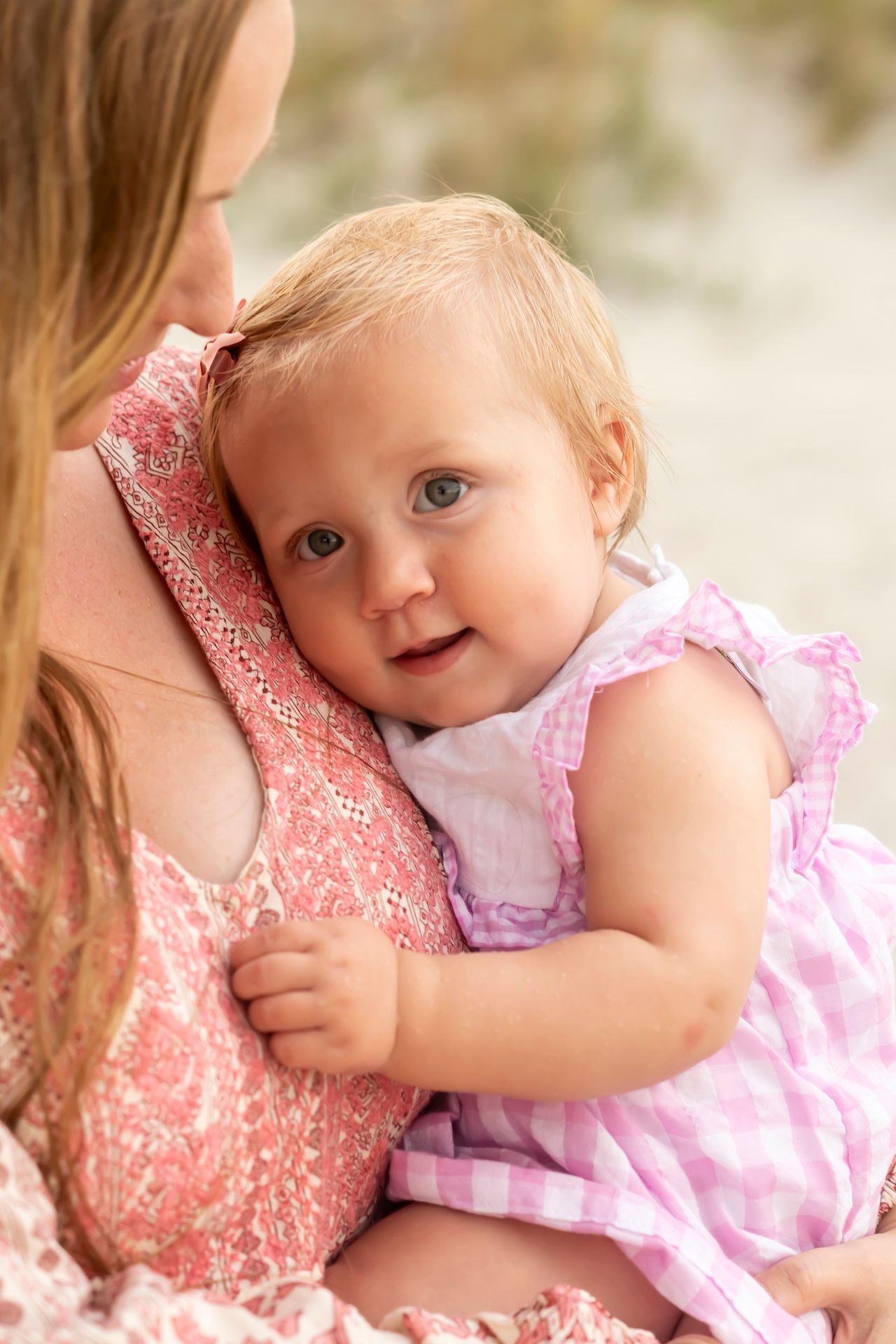 A baby in a pink and white dress snuggles with someone wearing coral lace in a soft outdoor portrait setting.