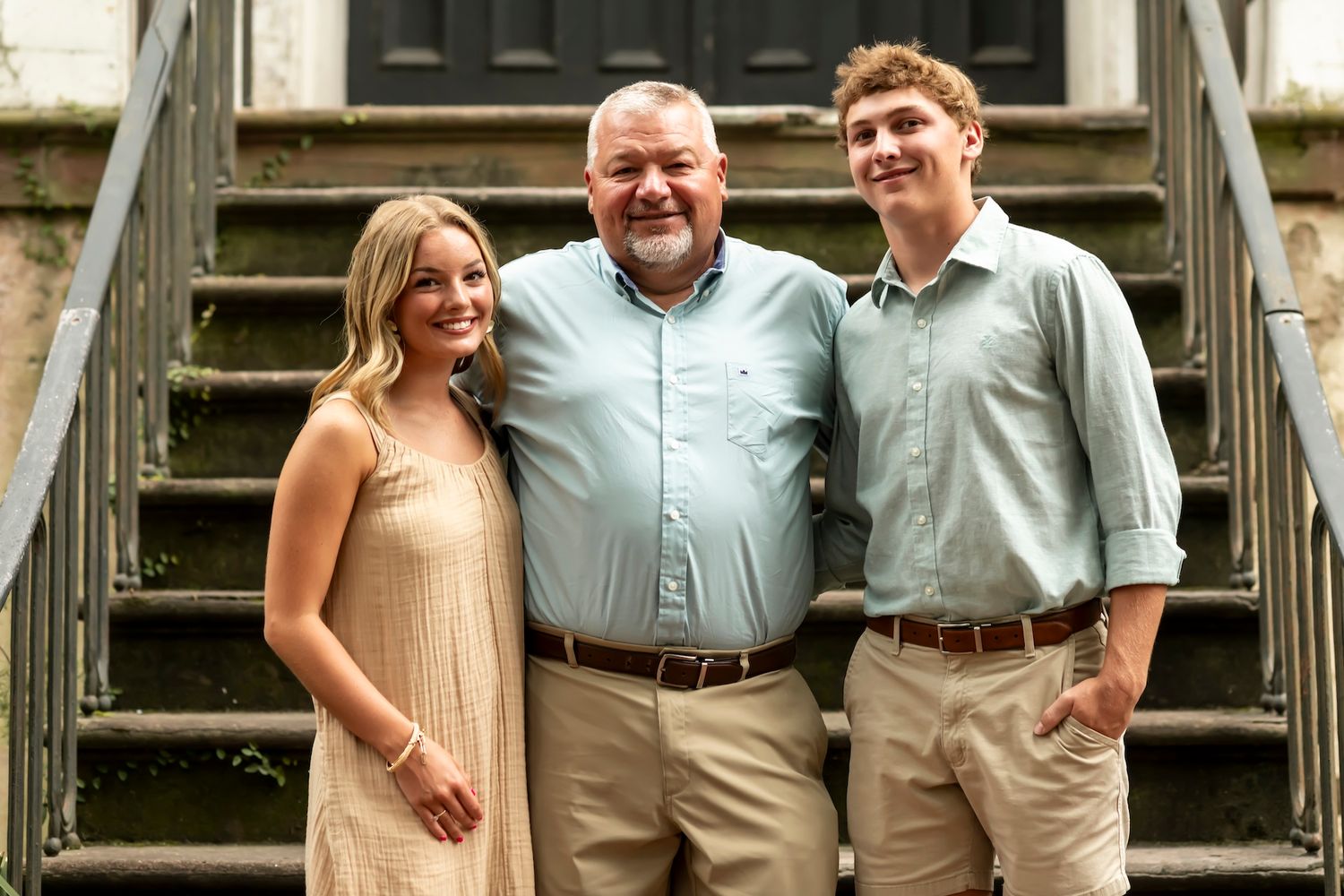 Three people stand together smiling on concrete steps in front of a building on a sunny day.