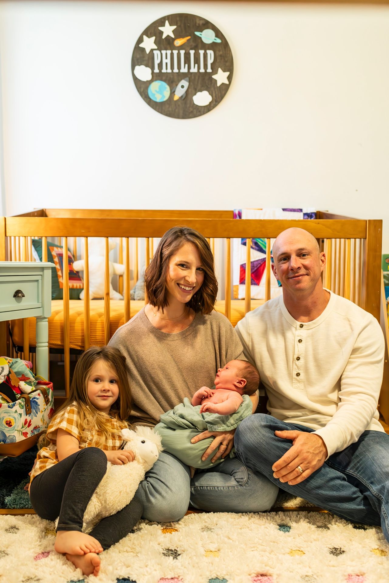 Family sits together on carpet near wooden crib with decorative name circle on wall showing stars and clouds.
