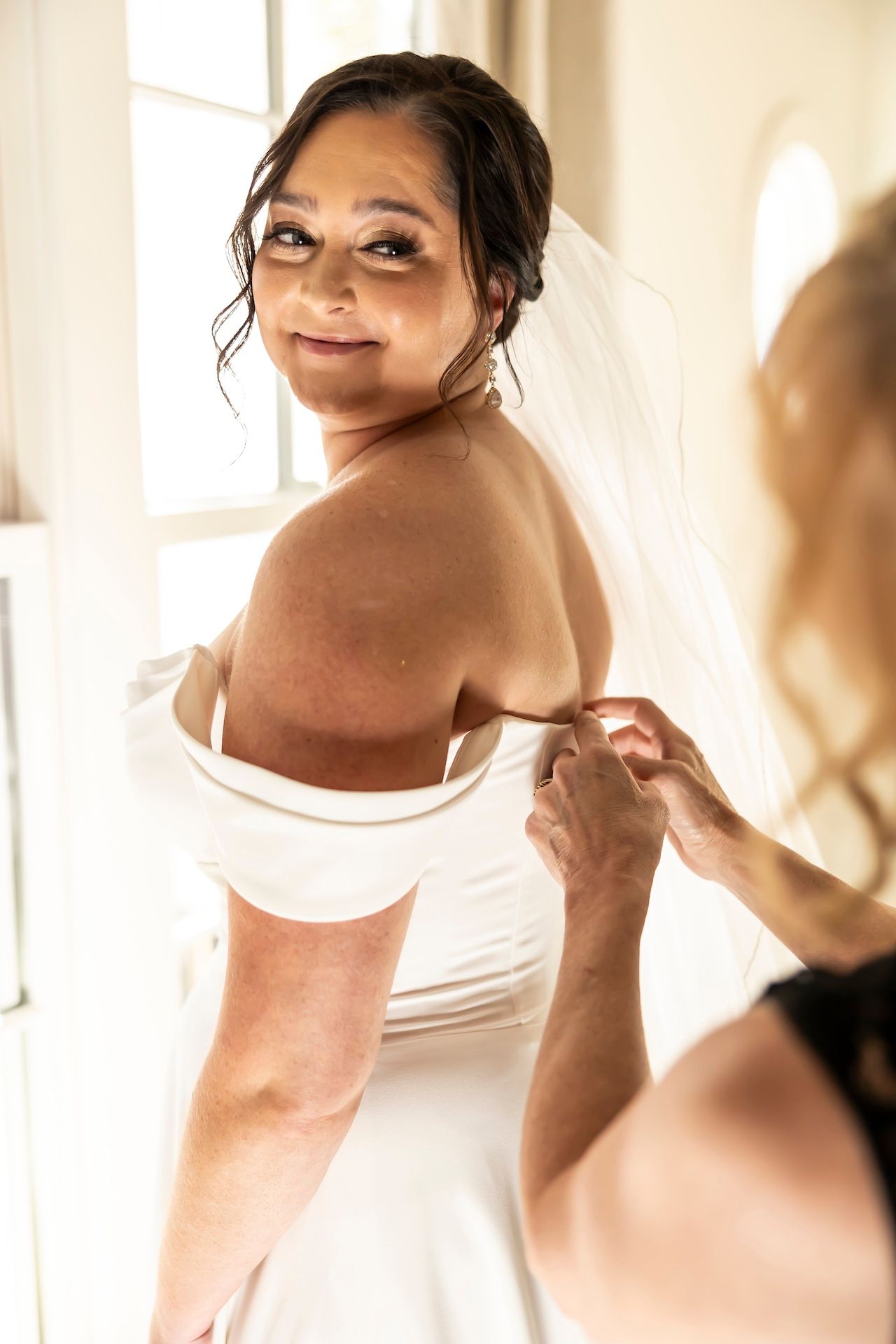 Someone getting help putting on a white wedding dress in warm natural lighting from a window.
