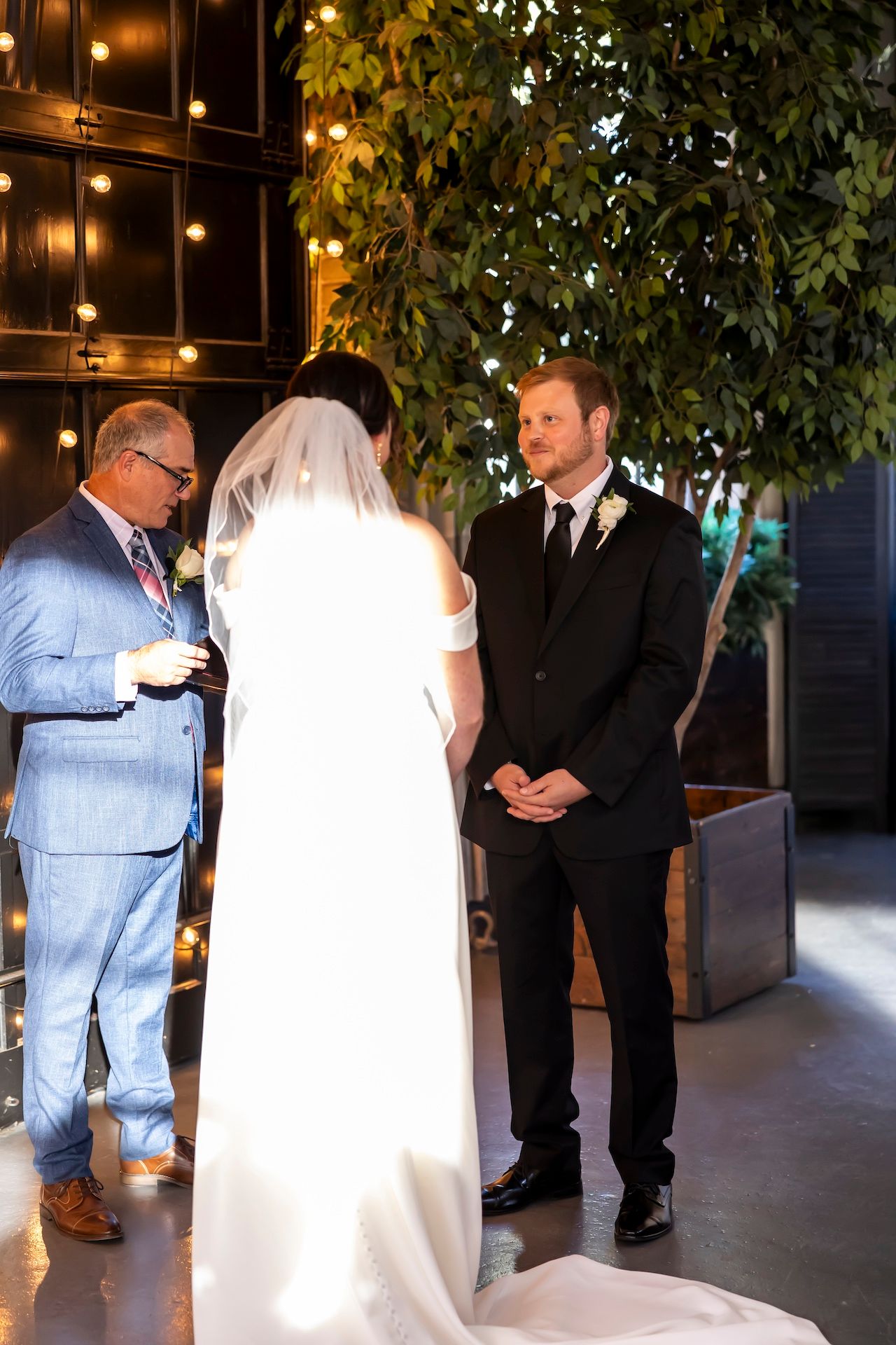 A wedding ceremony takes place in an intimate indoor venue with string lights and greenery decorations.