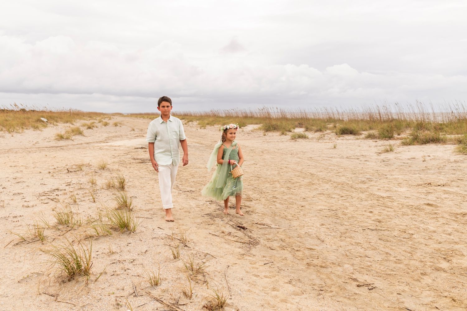 A person and a small child walk along a sandy beach with tall grass and dunes in the background.