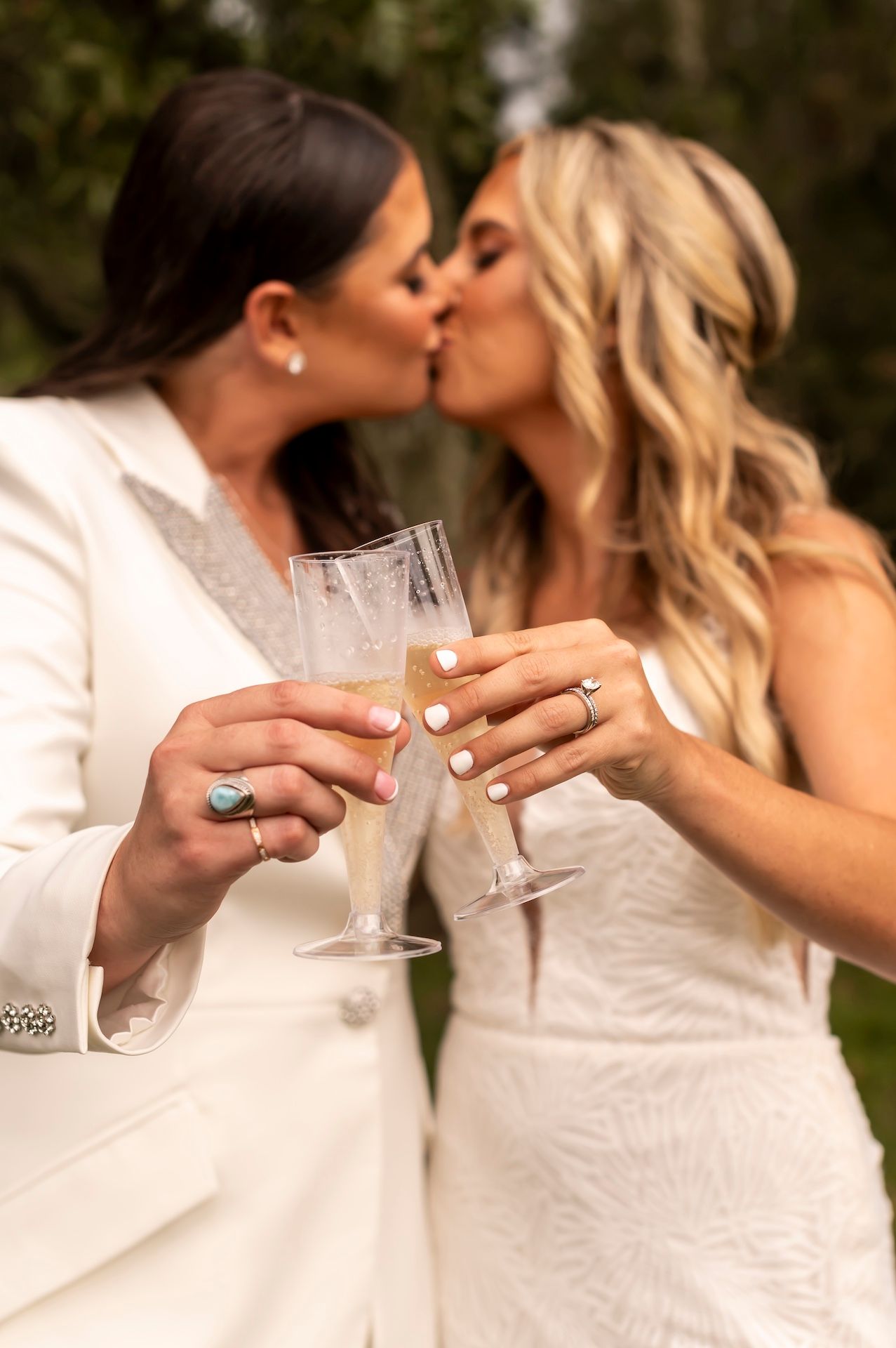 Two people in white formal attire share an intimate kiss while holding champagne glasses at their wedding celebration.