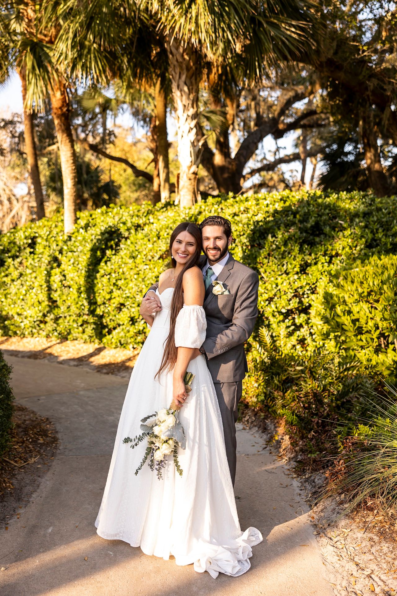 Newlywed couple poses on garden path surrounded by palm trees and lush hedges at sunset.