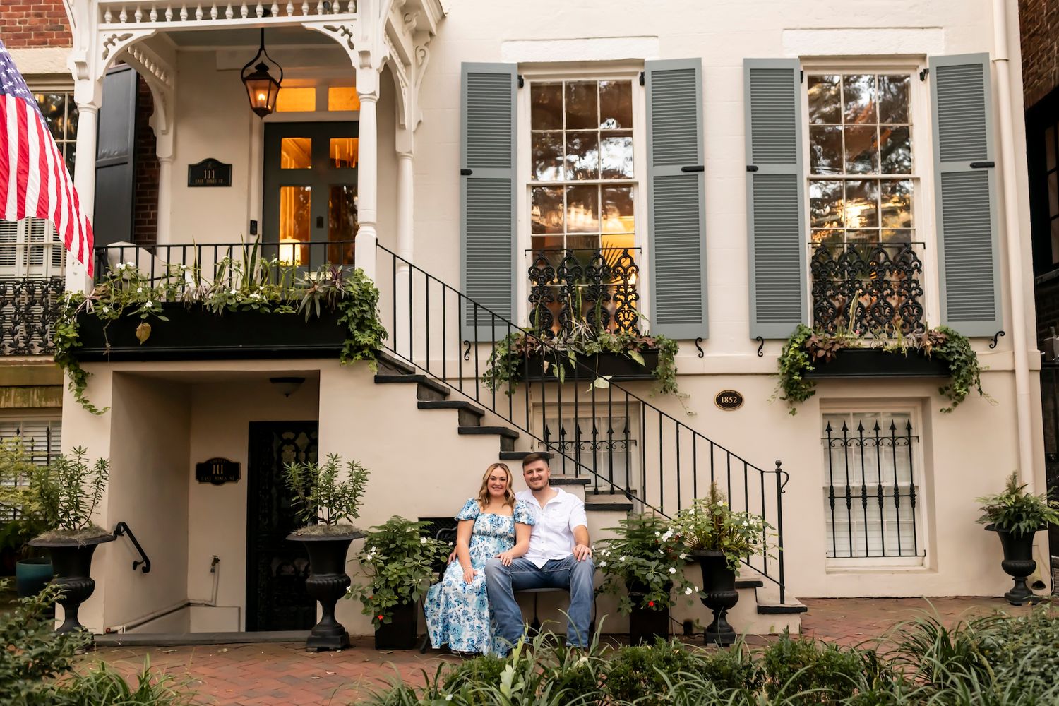 A couple sits on steps in front of a charming Charleston-style home with blue shutters and window boxes.