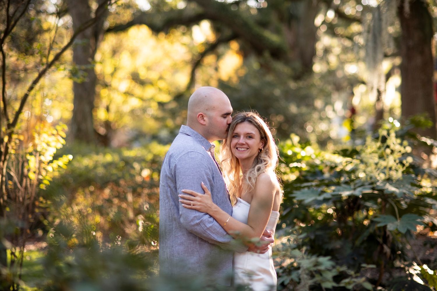 A couple embraces outdoors during a romantic photoshoot in a sun-dappled wooded setting at golden hour.