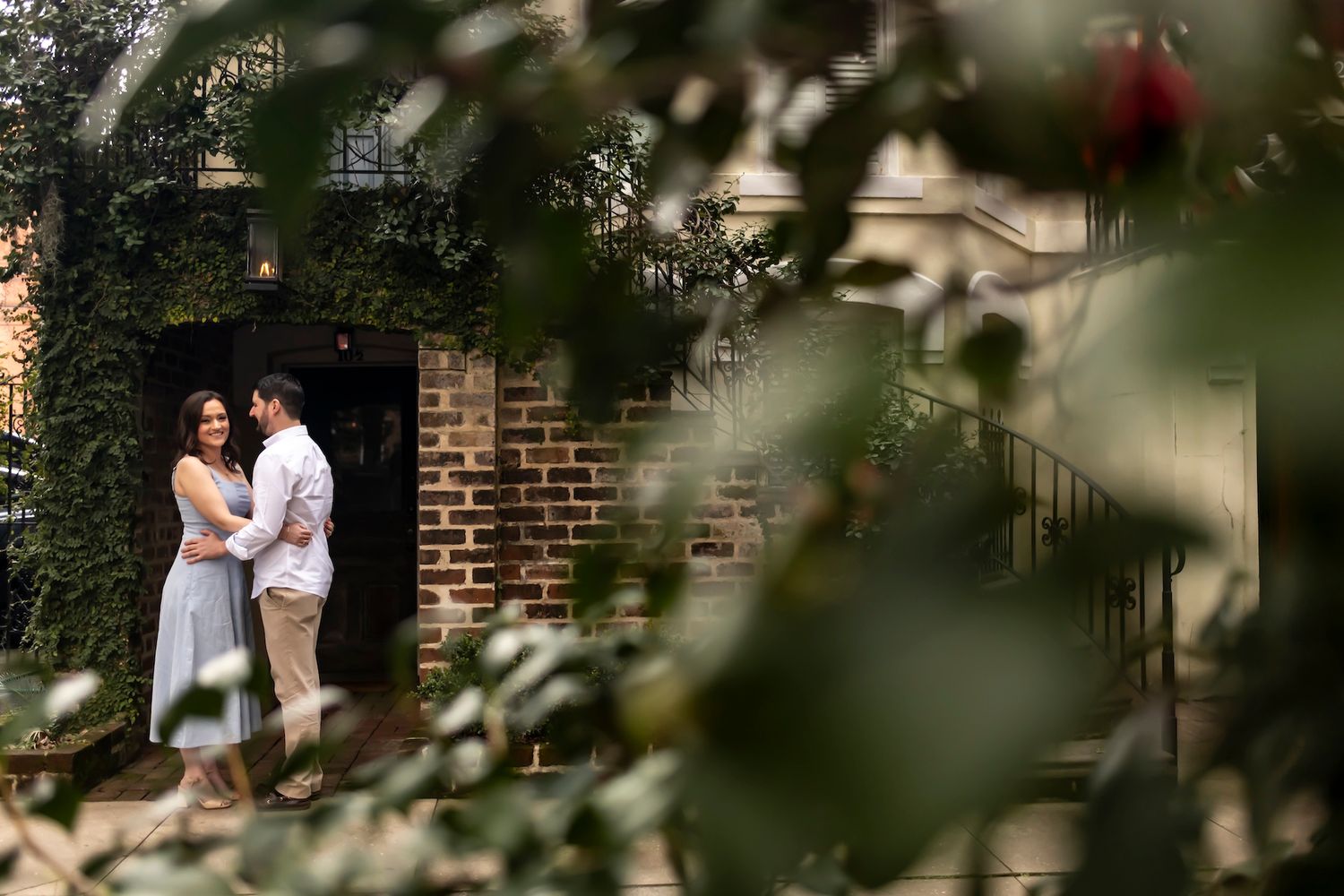 Two people in light clothing stand together in an outdoor brick archway framed by blurred foliage in the foreground.