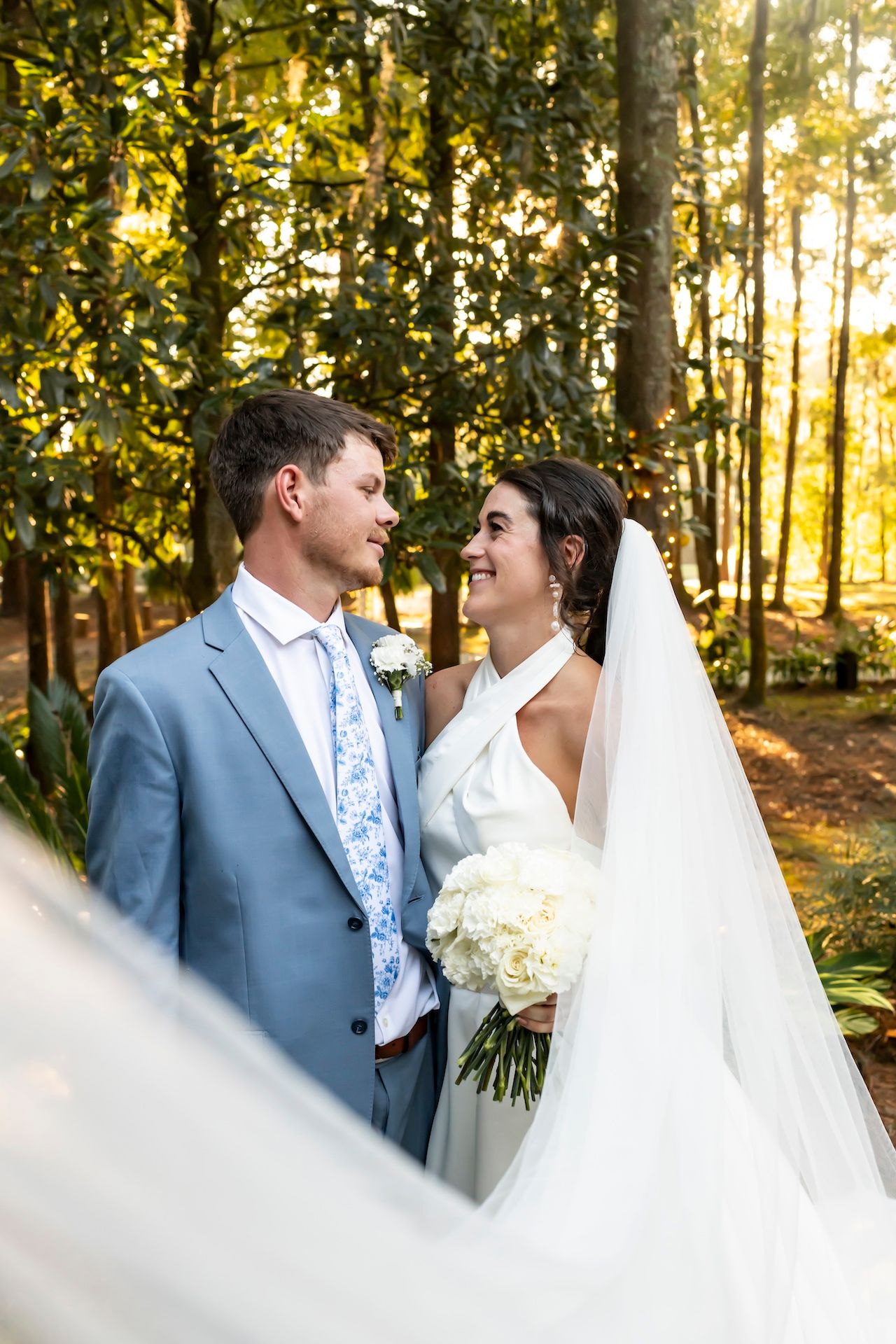 A joyful wedding couple shares an intimate moment in a sunlit forest with golden light filtering through autumn trees.