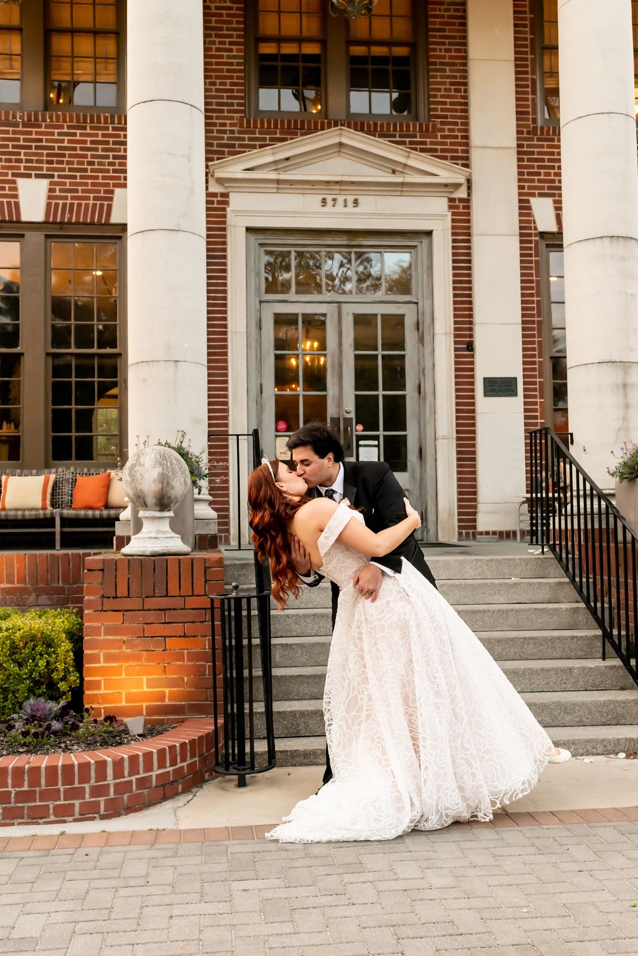 Romantic wedding couple shares a dip kiss on the steps of an elegant brick building with white columns and ornate entrance.