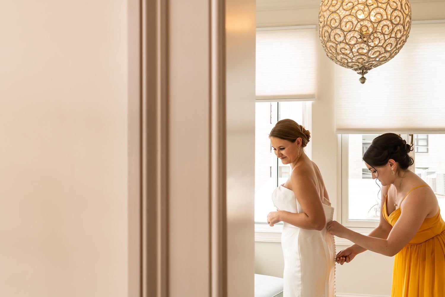 Two people help with preparations in a light-filled room with an elegant hanging light fixture.