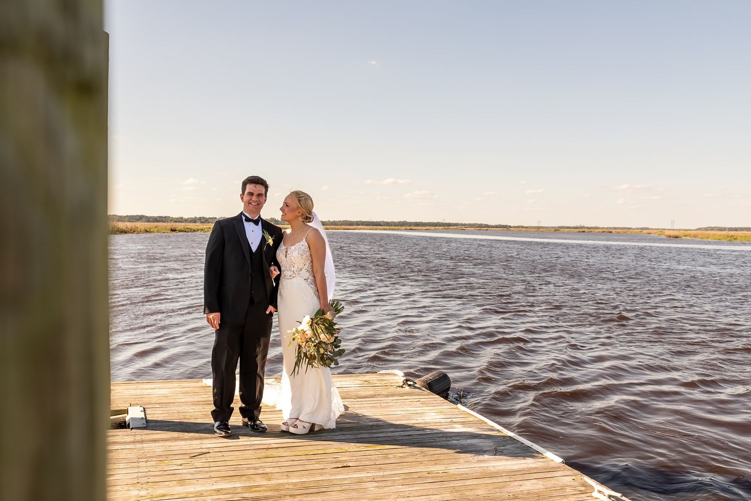Newlyweds stand together on a coastal dock with water and marshland in the background during golden hour.