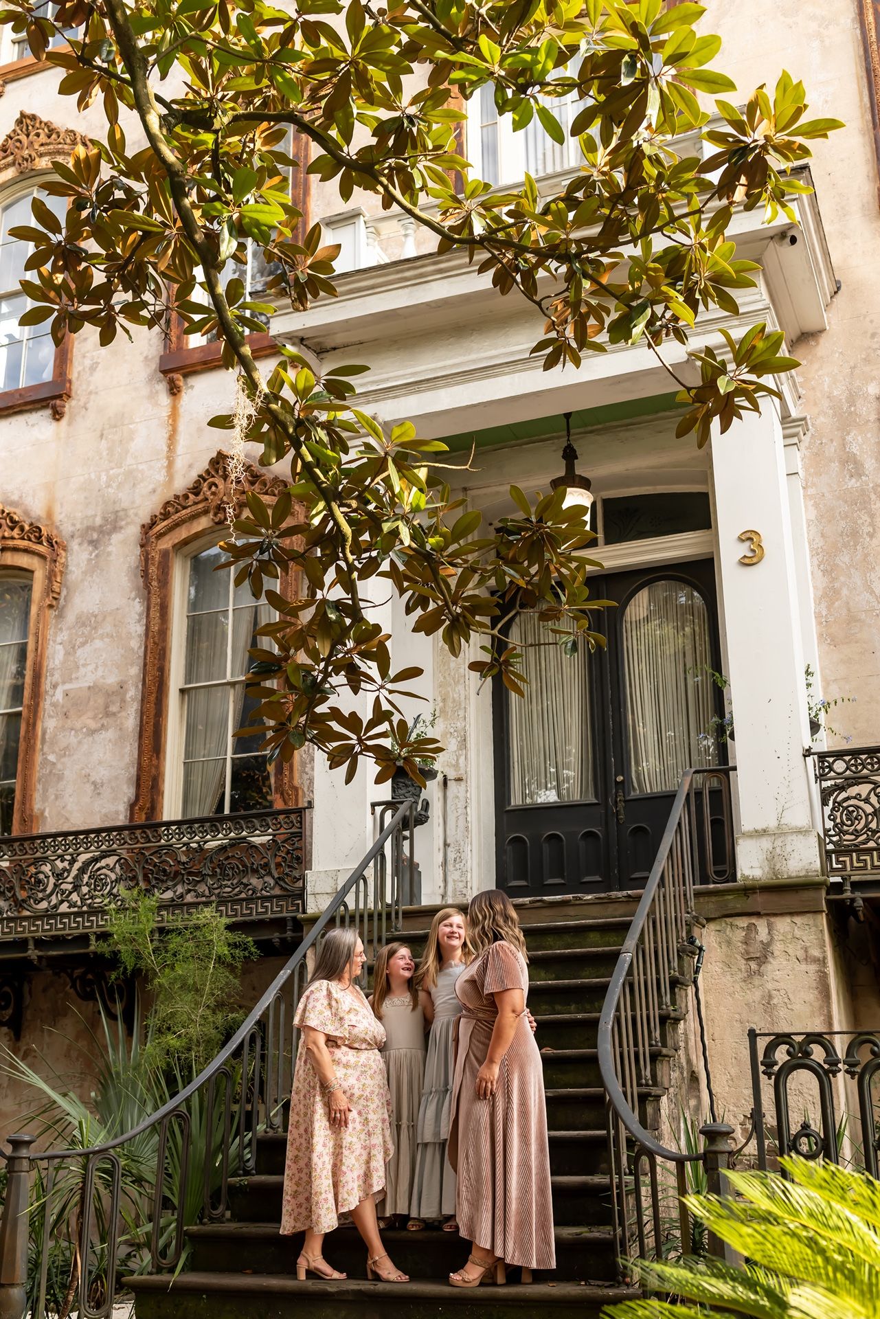 People in pink dresses gather under magnolia trees outside a historic Southern mansion with wrought iron details.