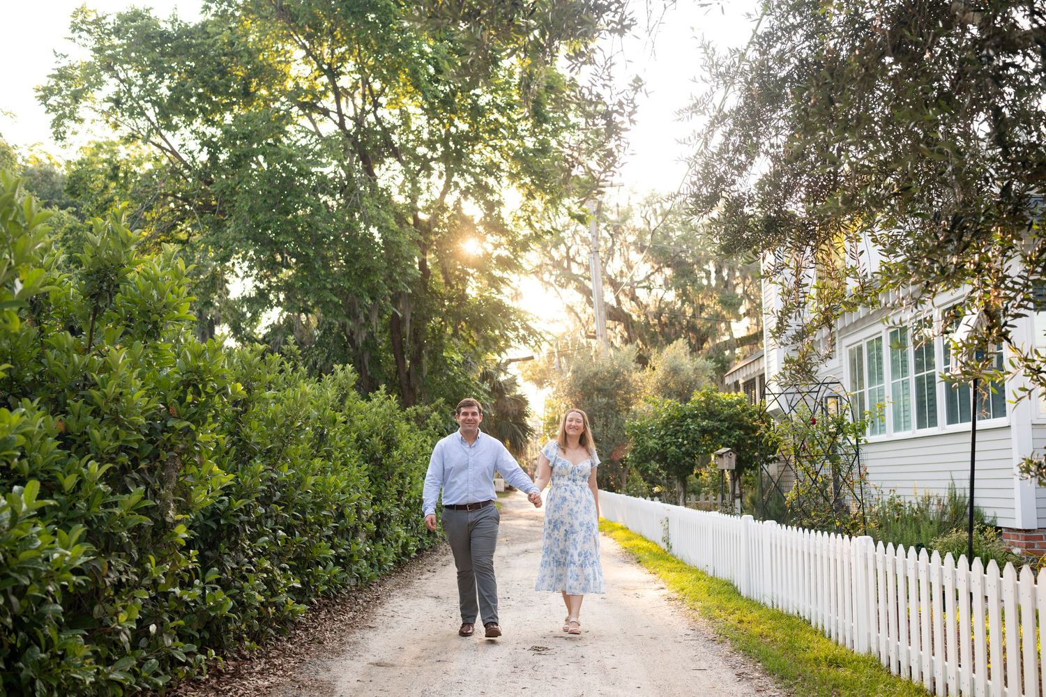 A couple walks down a sunlit path bordered by lush greenery and a white picket fence on a warm afternoon.