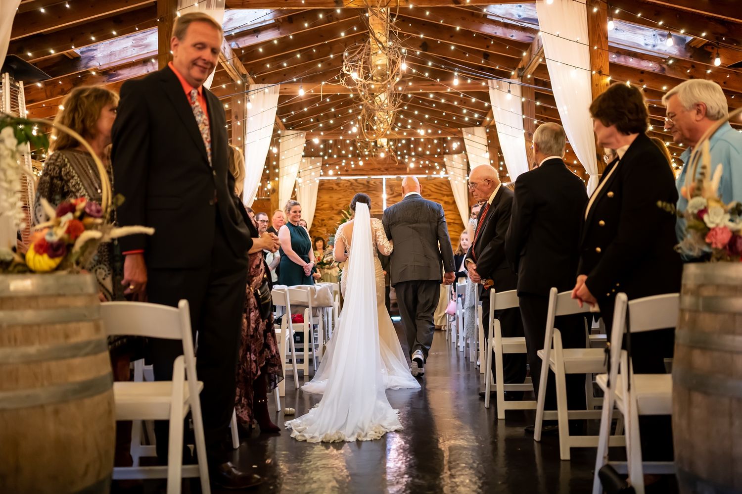 Wedding ceremony in rustic barn venue with glowing lights and wine barrels lining aisle as guests look on.