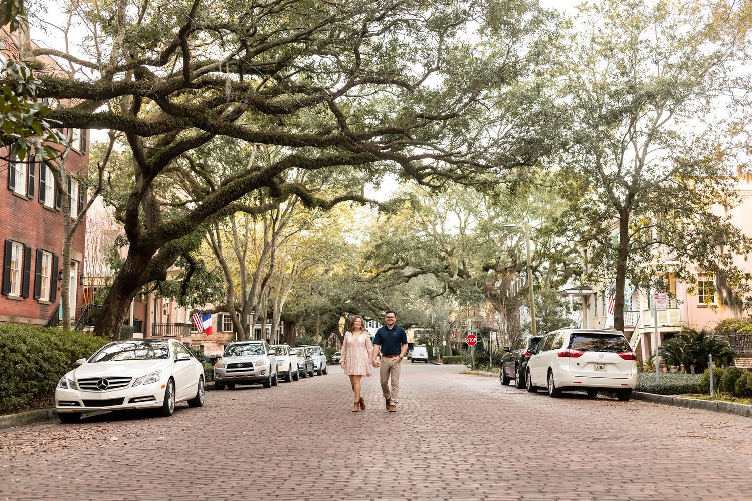 Two people stroll down historic brick street lined with parked cars and moss-draped oak trees in downtown Charleston.