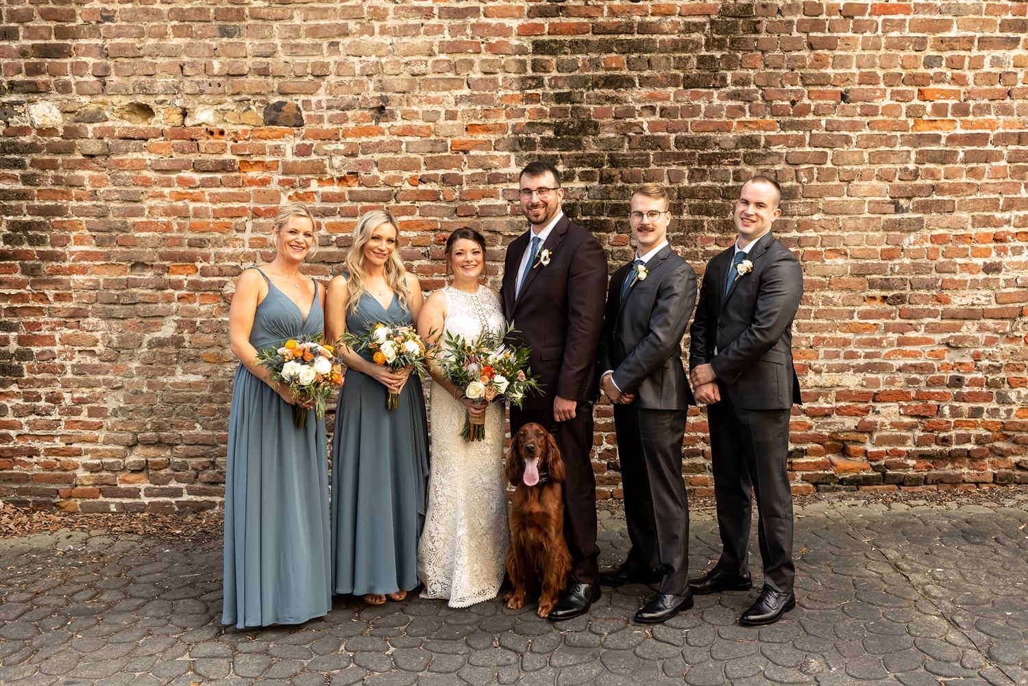 A wedding party stands together in front of a rustic brick wall wearing dusty blue dresses and dark suits.