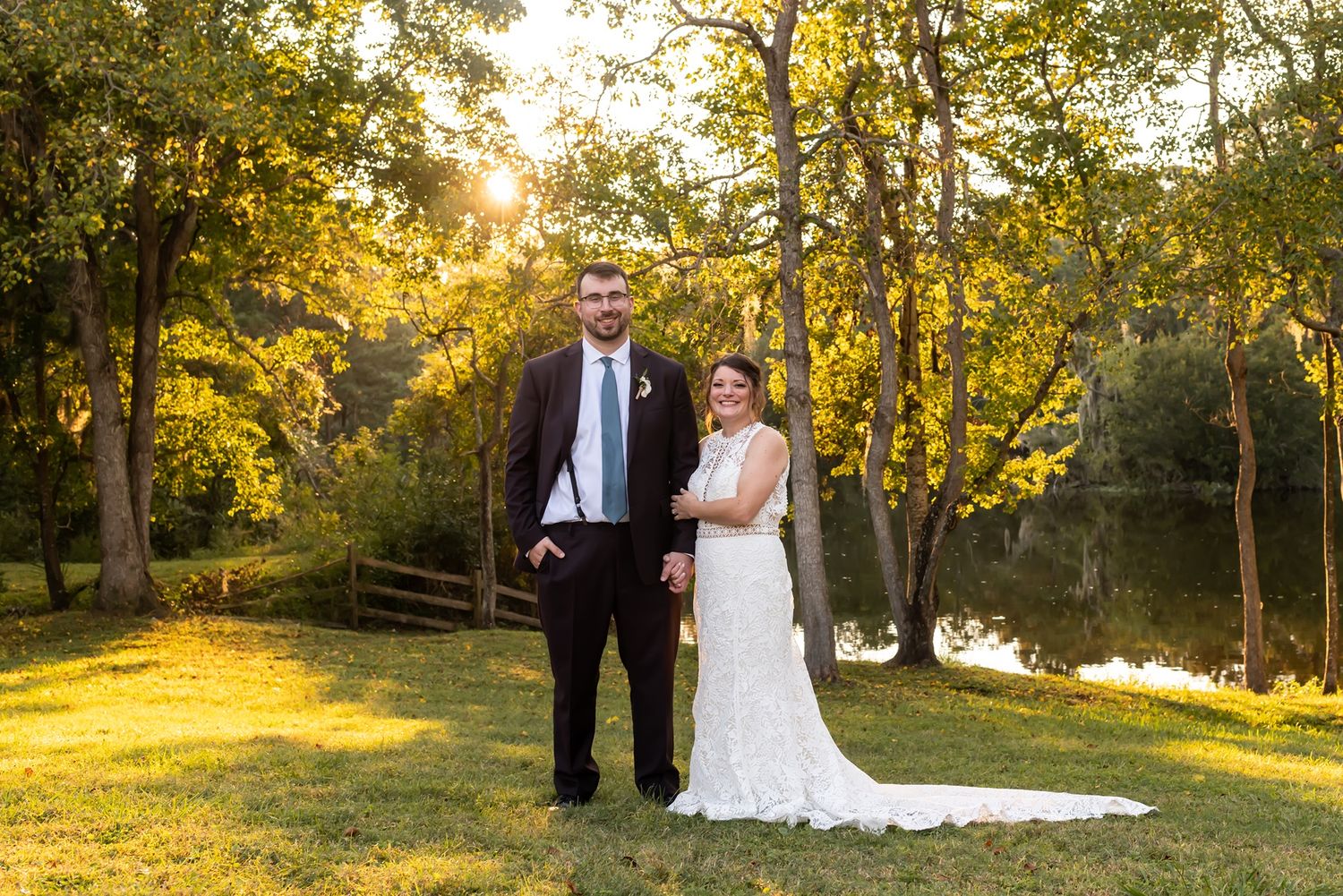 Newlywed couple stands together in golden sunlight under autumn trees by a tranquil pond at sunset.