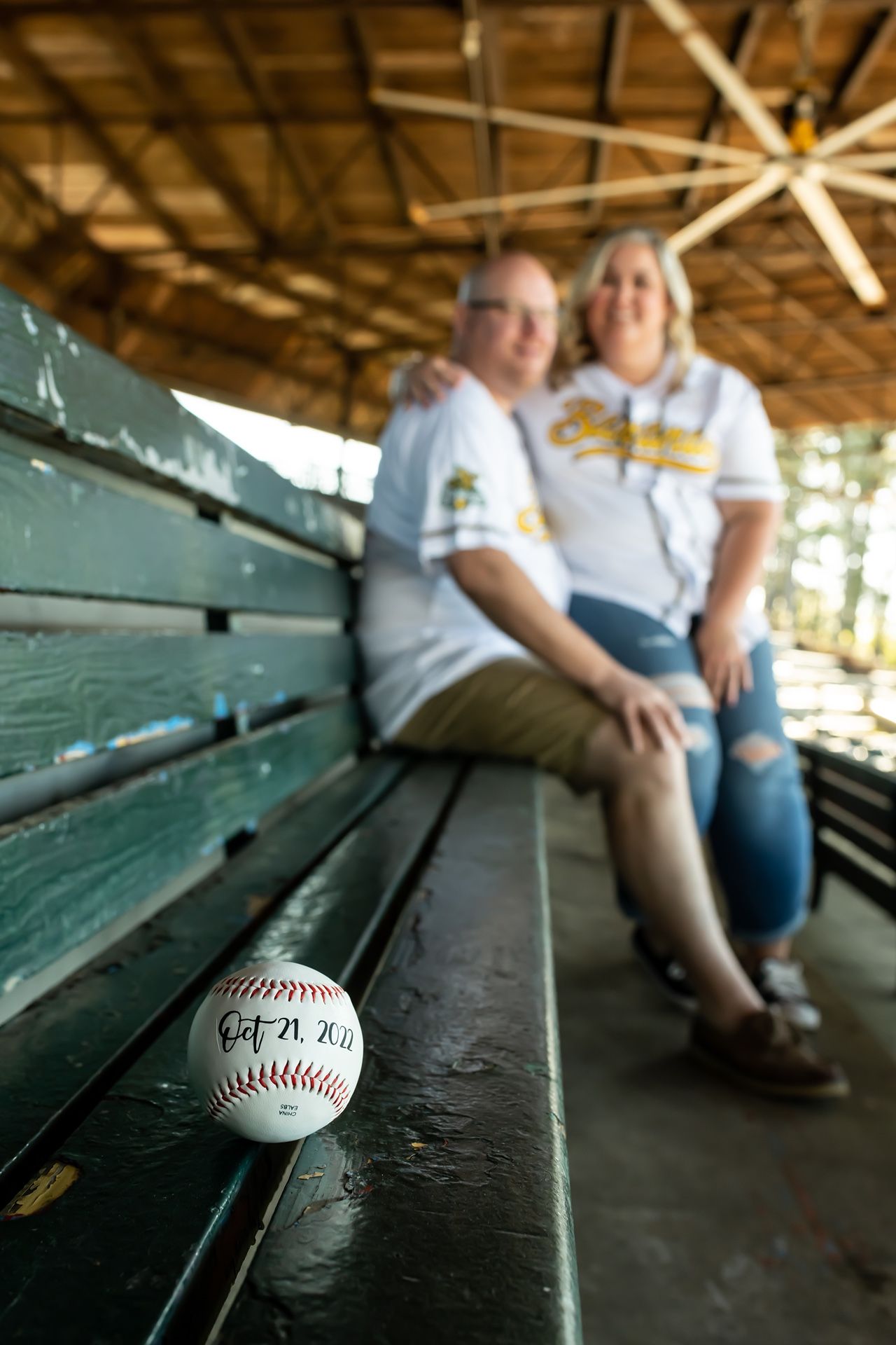 Couple in matching white shirts sitting on old green bleachers with a baseball in the foreground.