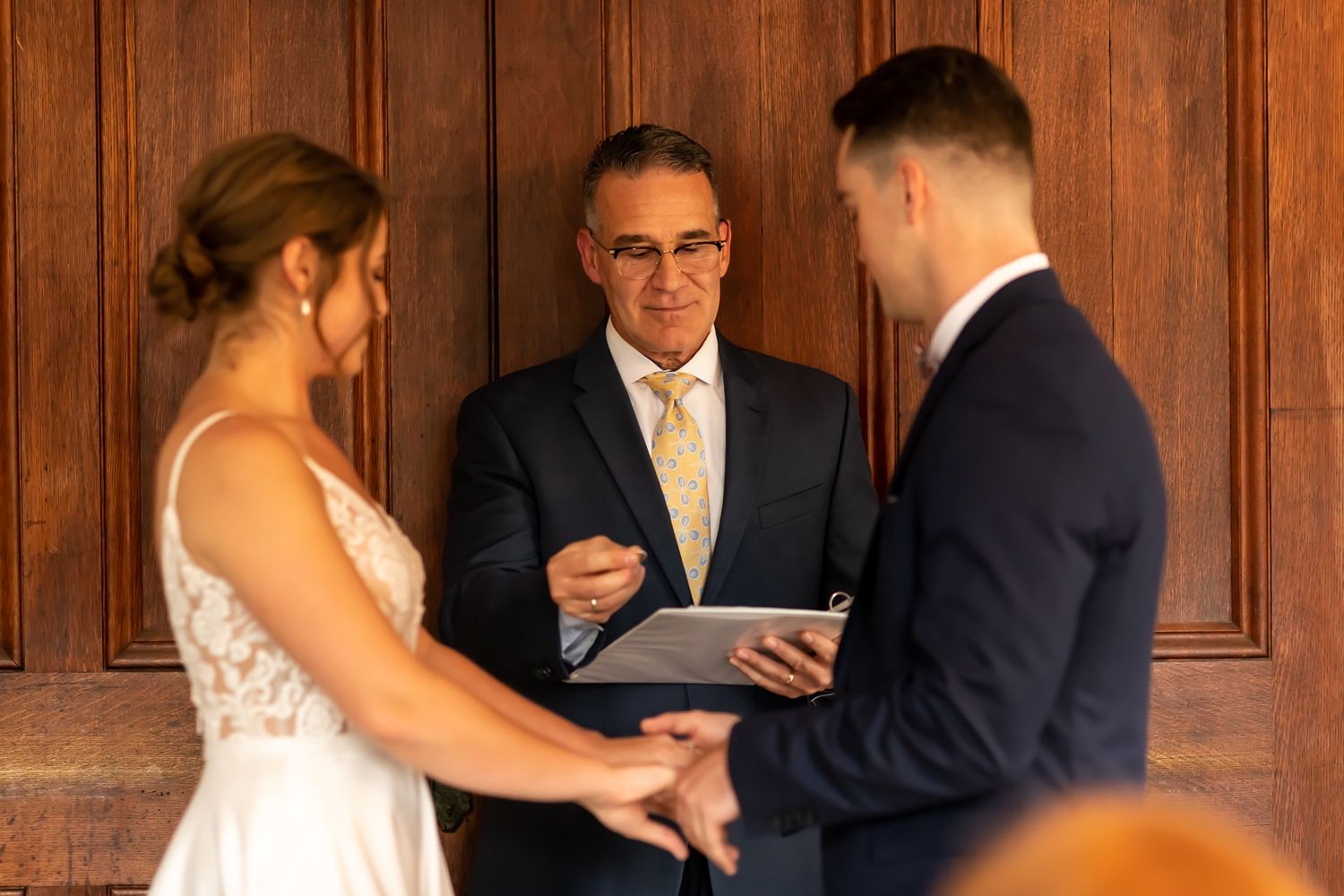 A couple exchanges vows during a ceremony while an officiant reads from a tablet in front of wood paneled walls.
