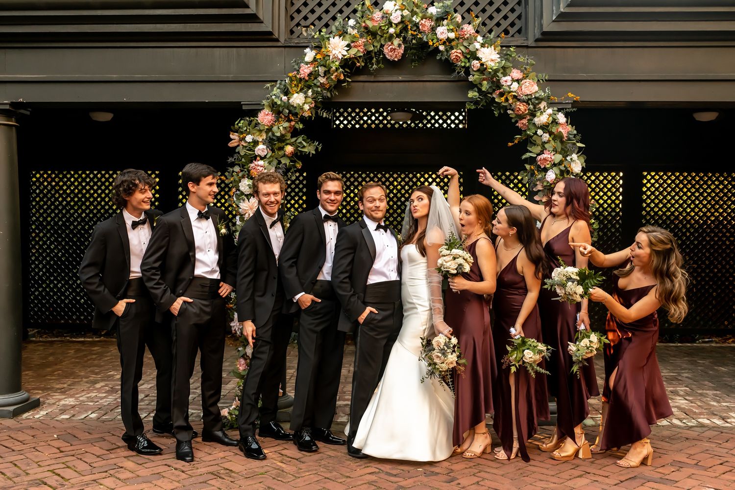 Wedding party poses together at night on a brick patio with string lights and floral decorations overhead.