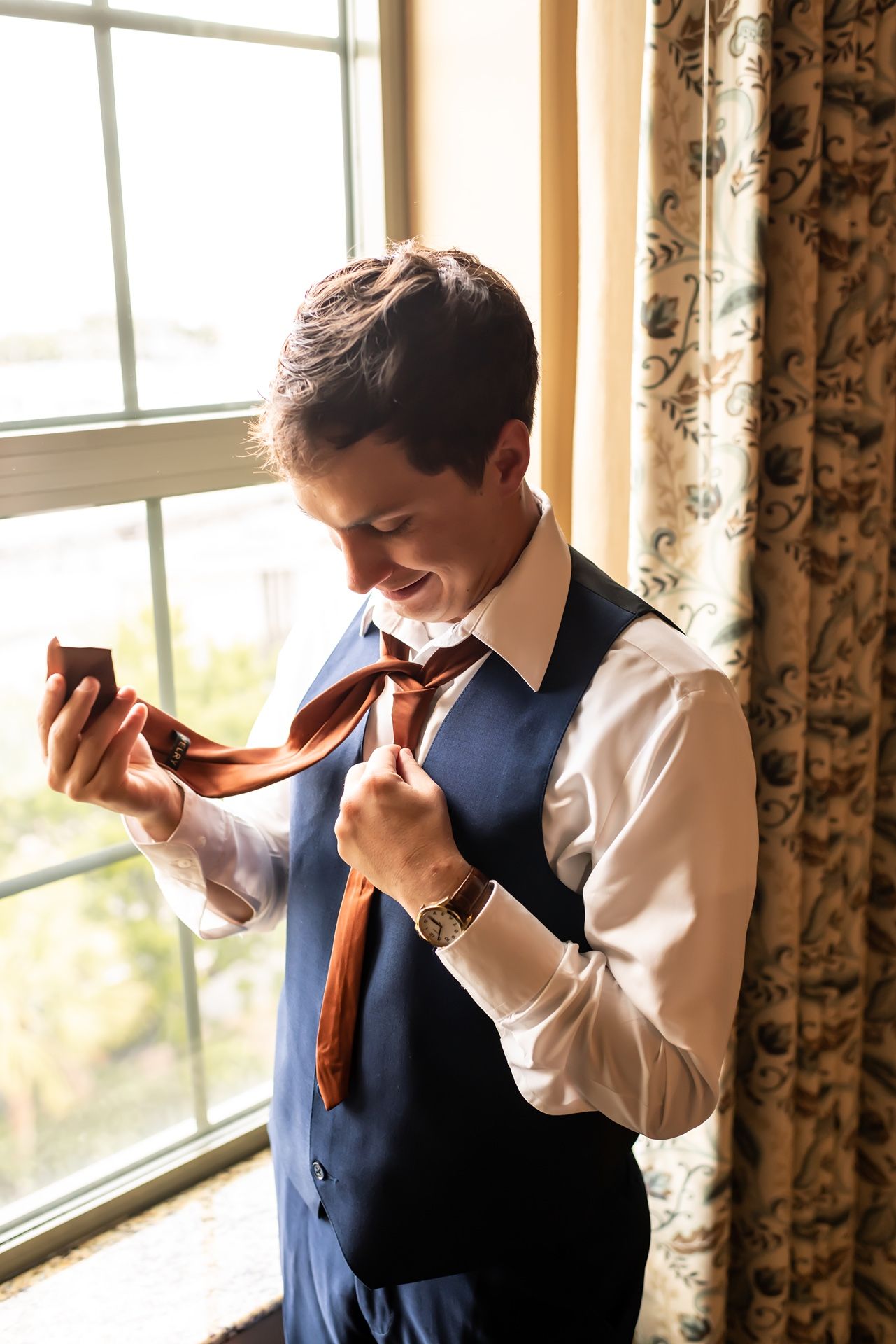 Person in navy vest and white shirt holding brown object while standing by window with floral curtains in warm lighting.