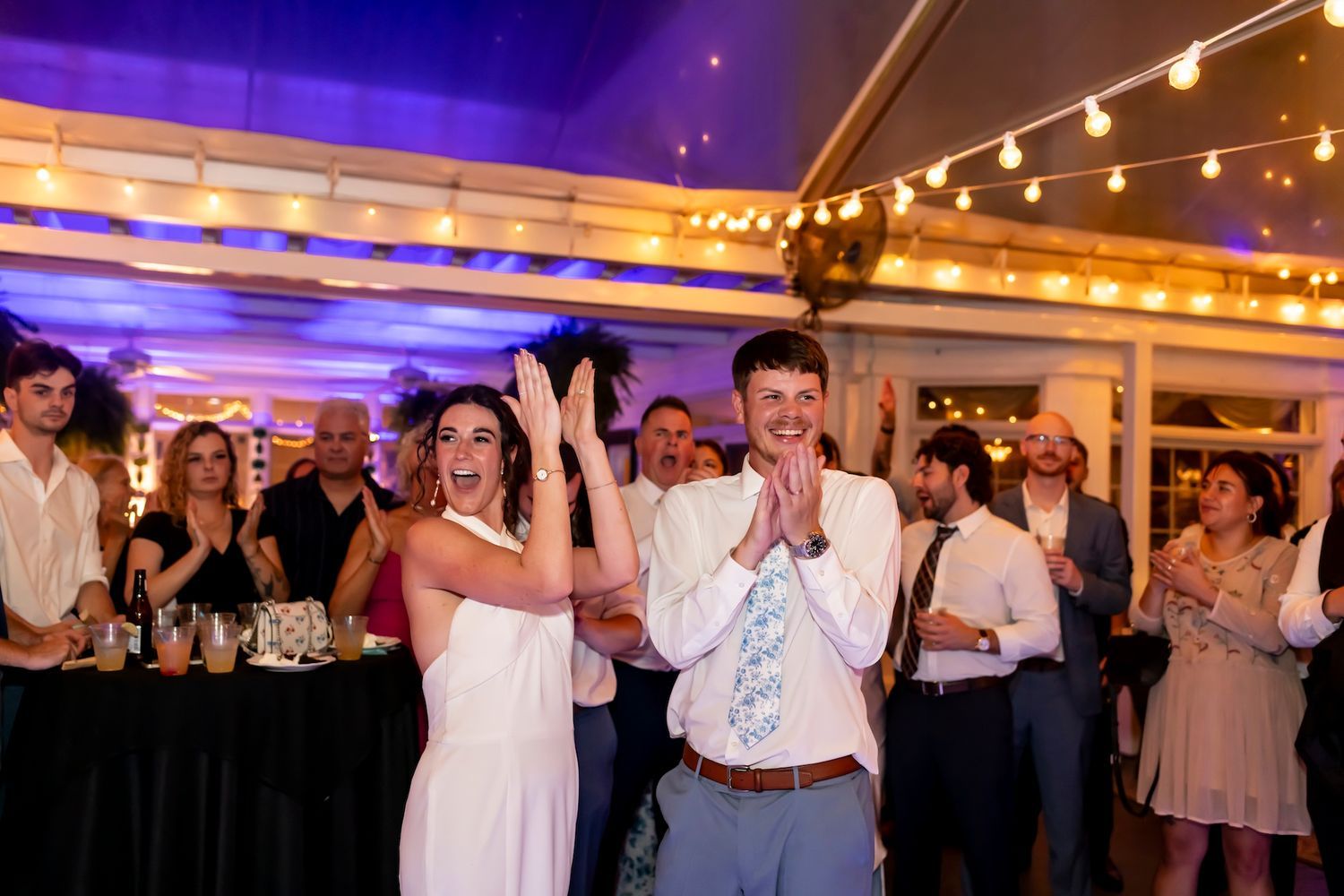 Wedding celebration with guests dancing and celebrating under purple lighting and string lights at an elegant reception venue.