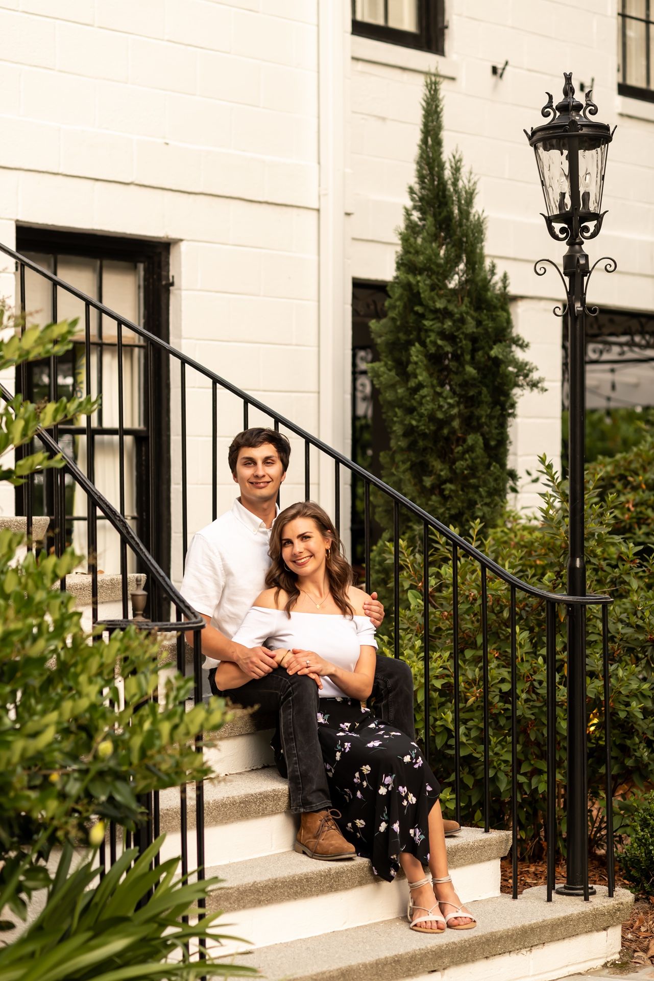 Two people share a romantic moment sitting on outdoor stone steps with ornate black iron railings and greenery.