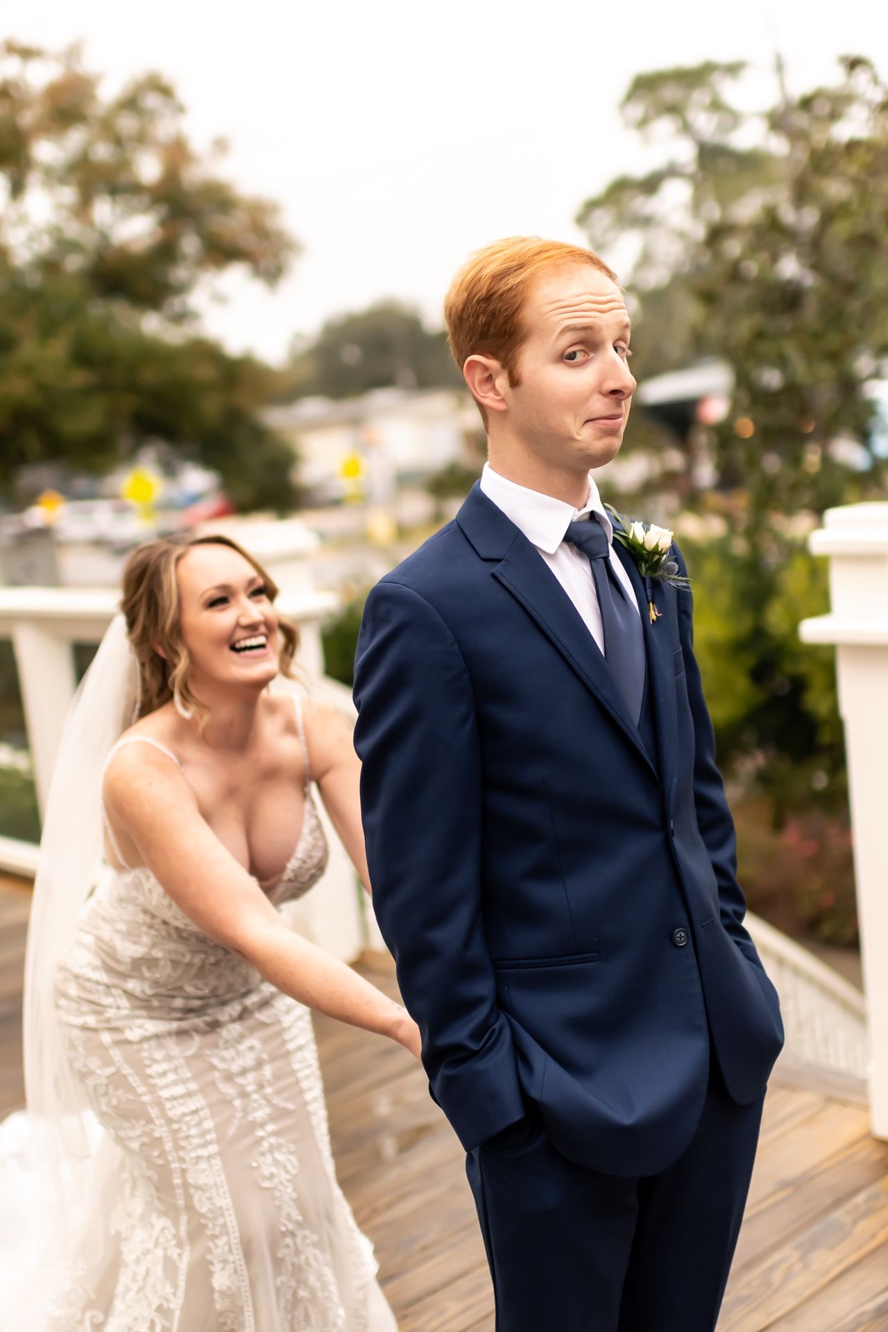 A candid moment as a bride in a fitted lace gown laughs behind her groom wearing a navy suit on their wedding day.