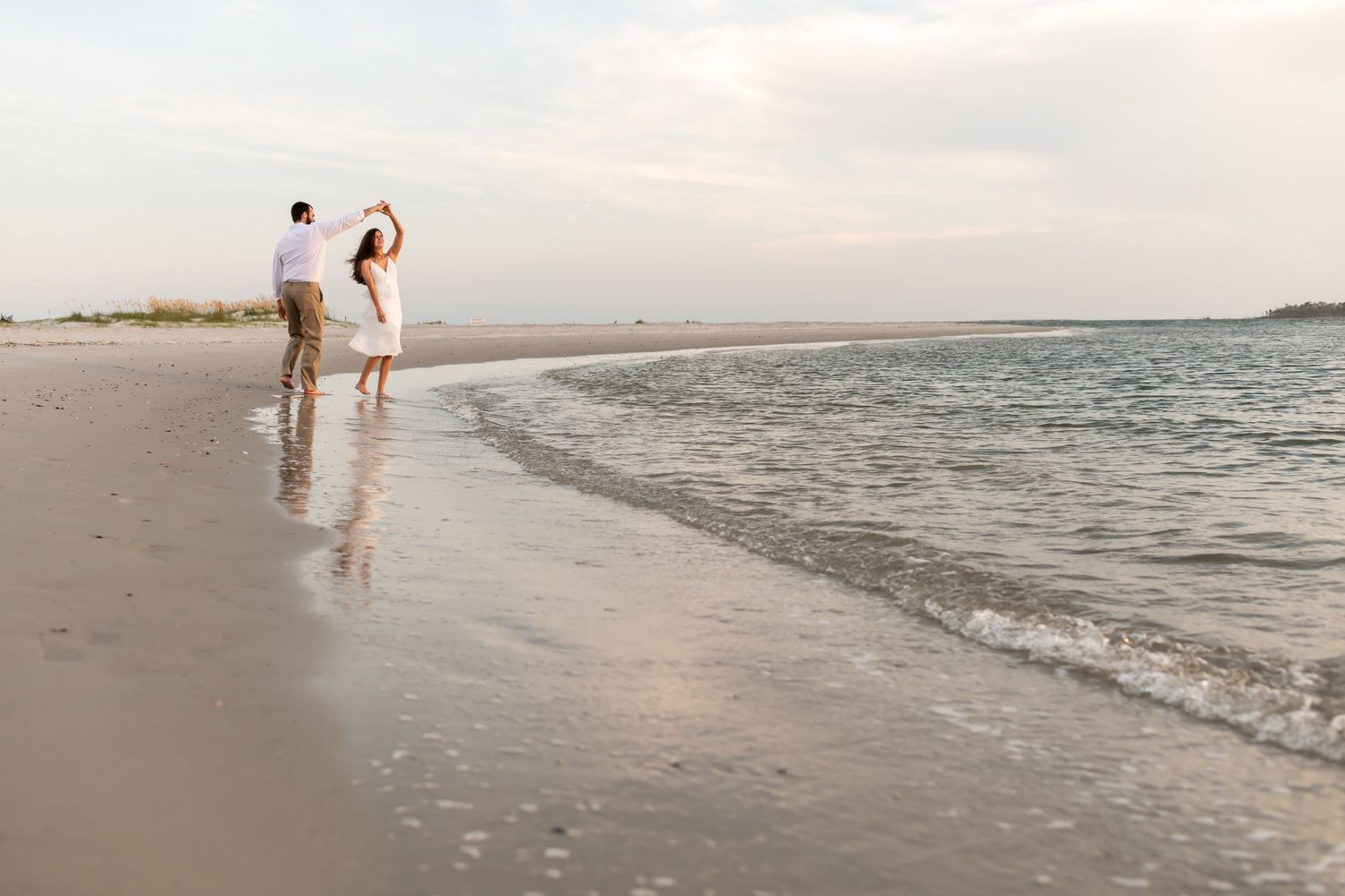 Two people walk along a misty beach shoreline during sunset with waves gently rolling onto the sand.