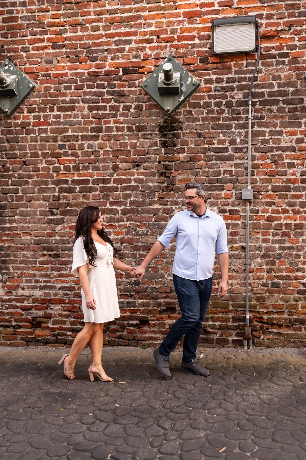 A couple strolling together against a vintage brick wall with industrial light fixtures in an urban setting.