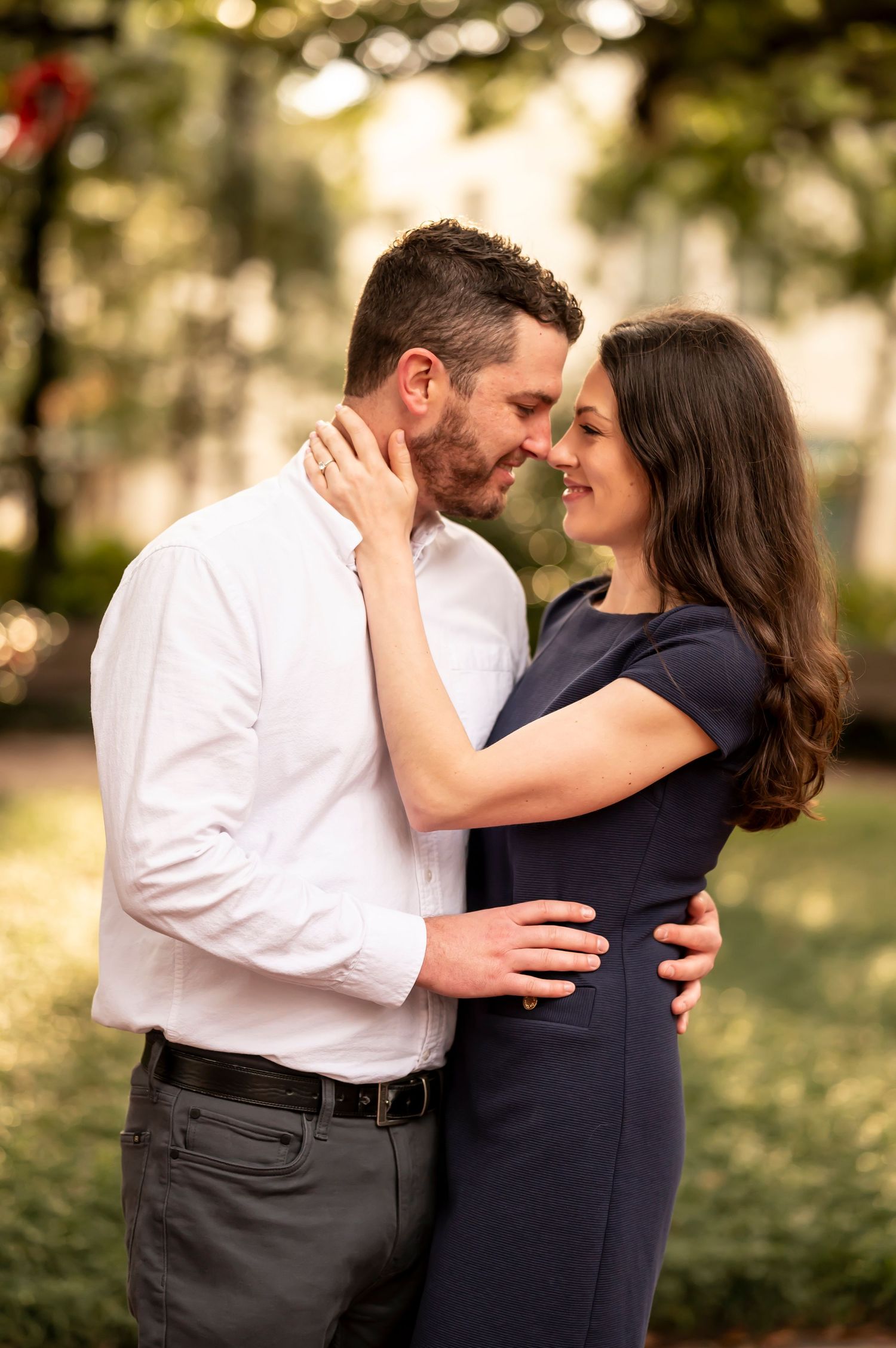 A couple shares intimate romantic moments in a park during a golden hour photo session with sunlit trees in background.