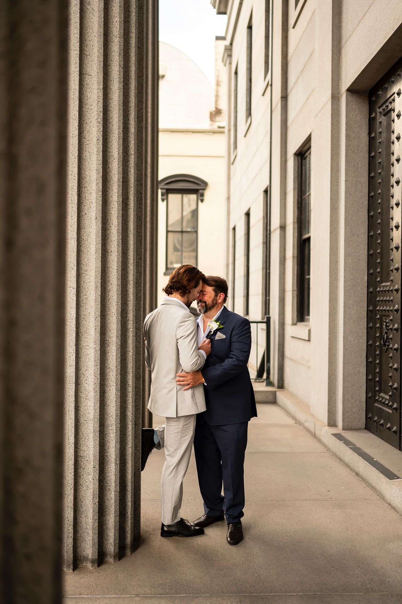 A couple in wedding attire share an intimate moment in an elegant urban setting with classic architecture.