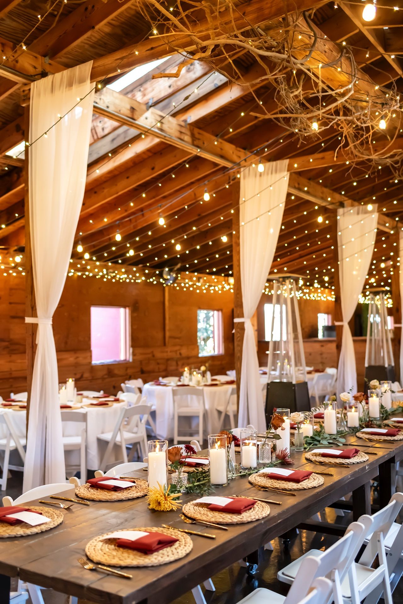 Long rustic wooden tables set for a wedding reception with white drapes and twinkling string lights in a barn venue.