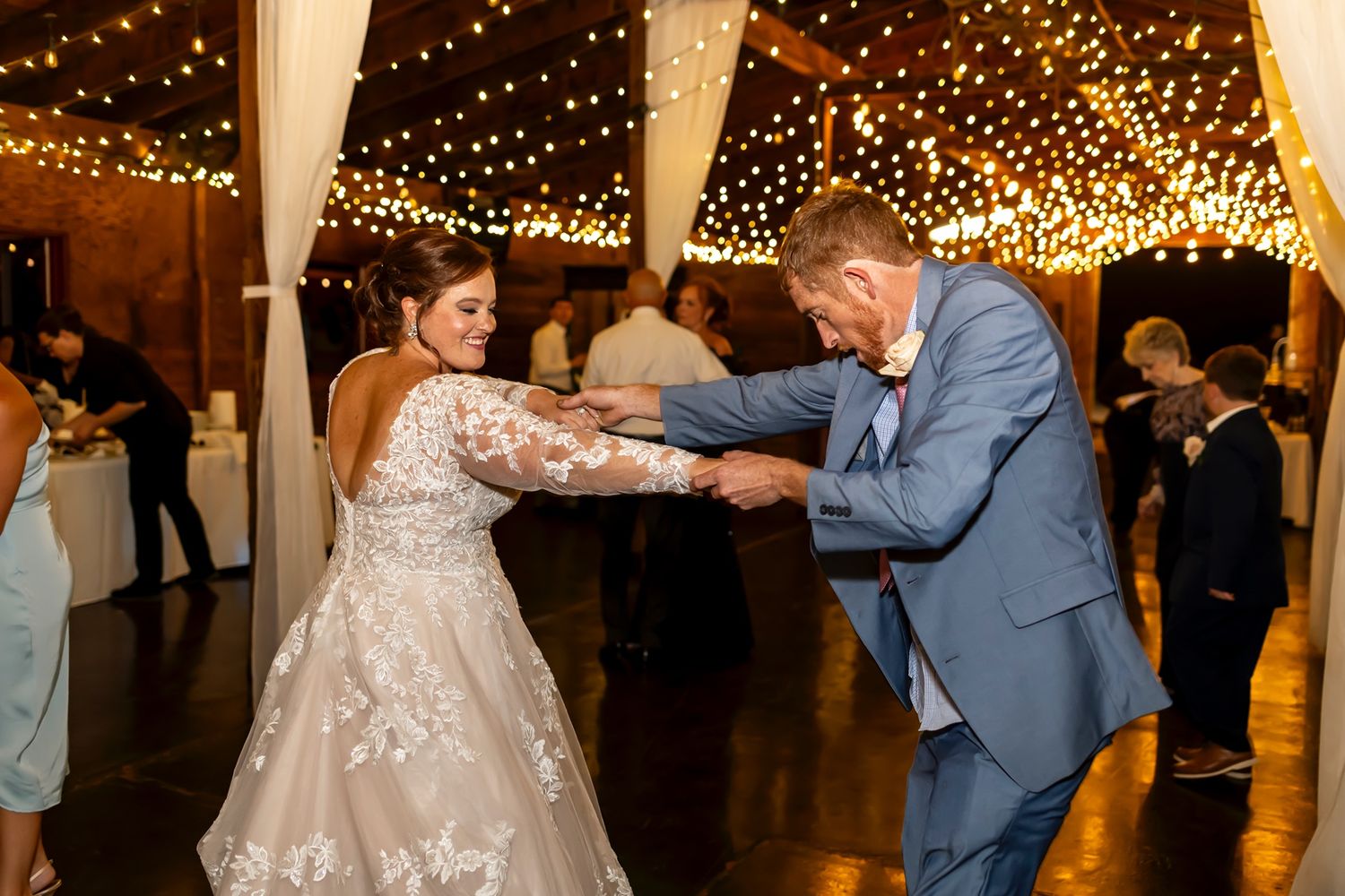 Two people dance together in an indoor reception venue decorated with string lights and white drapes overhead.