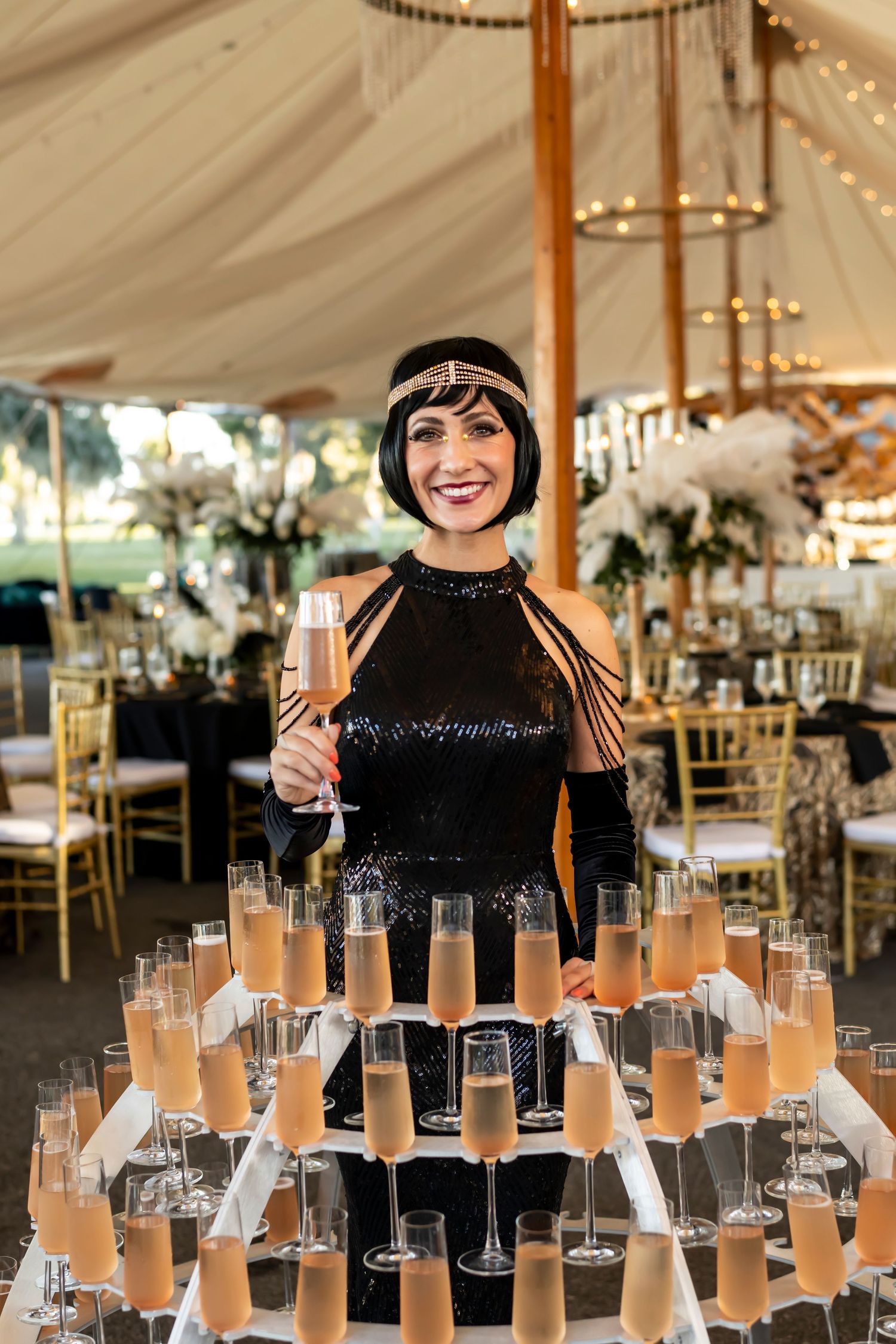 Row of champagne flutes arranged at an elegant wedding reception under a white tent with gold chairs and floral decor.