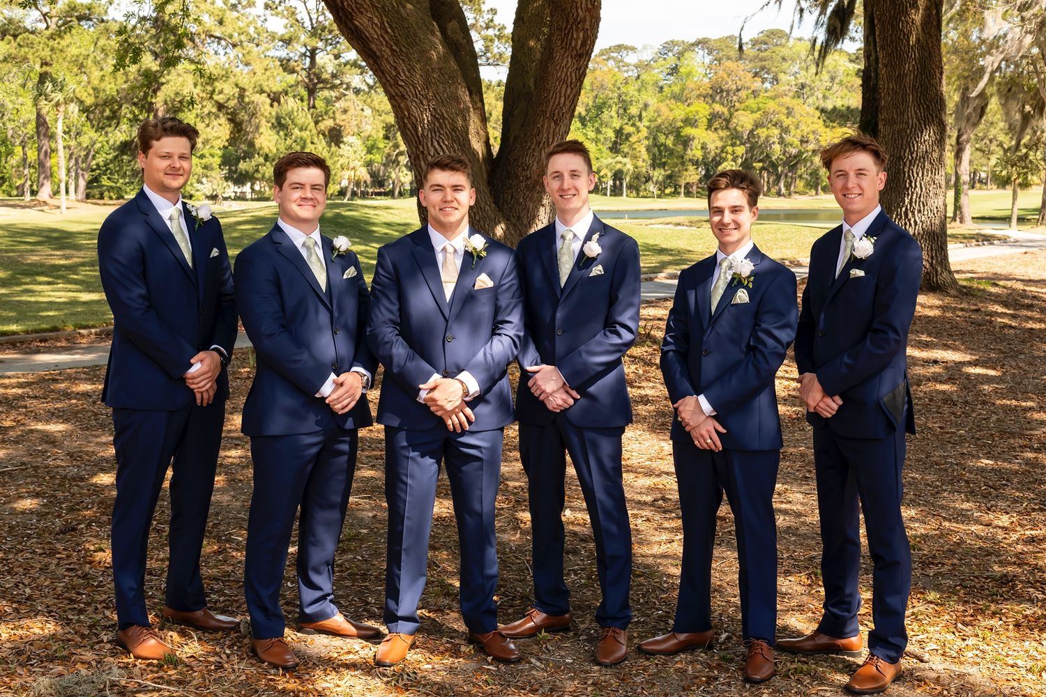 Groomsmen in matching navy suits and brown shoes pose together under a large tree in a park setting.