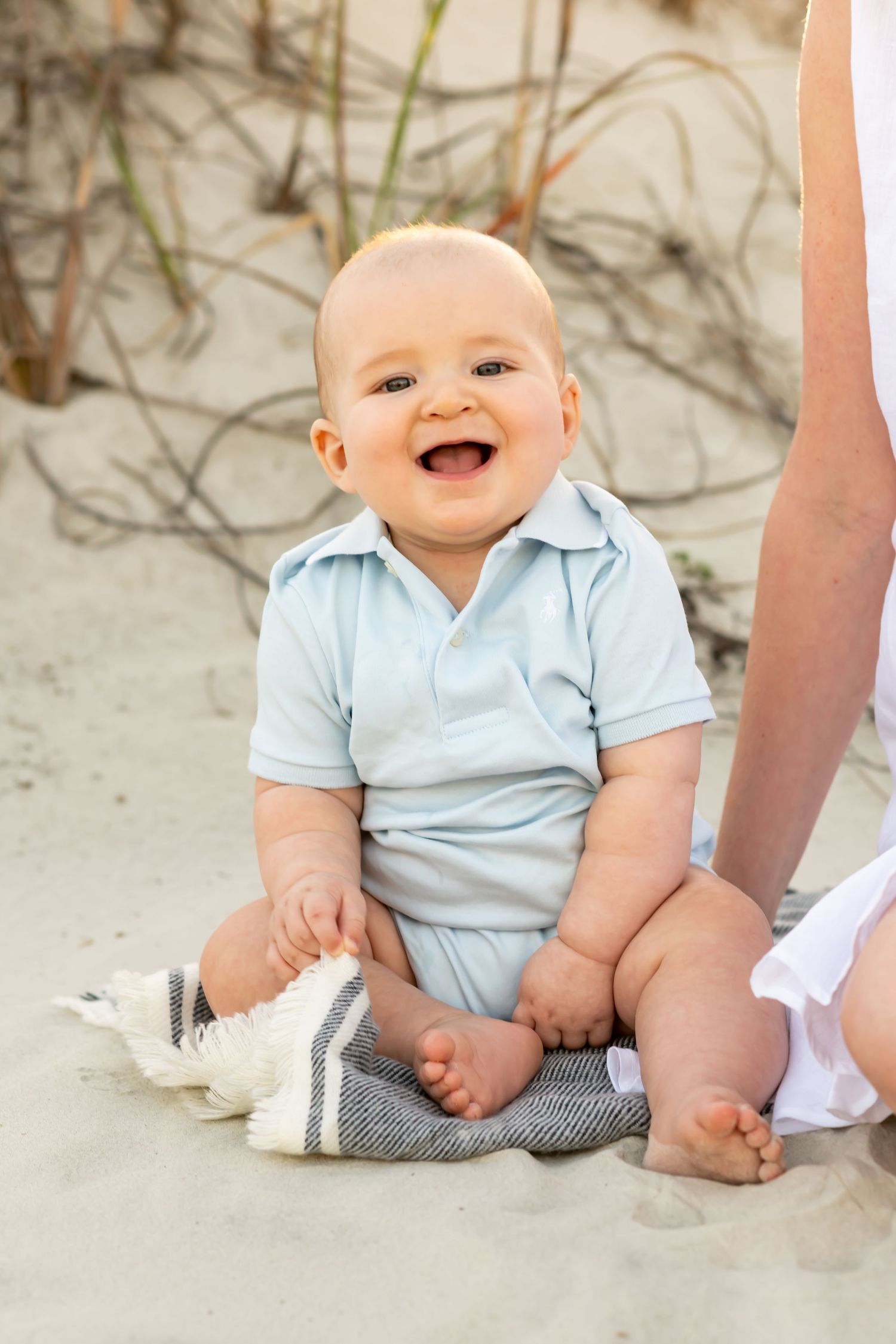 A happy baby in light blue outfit sits on sandy beach with sea oats in background.