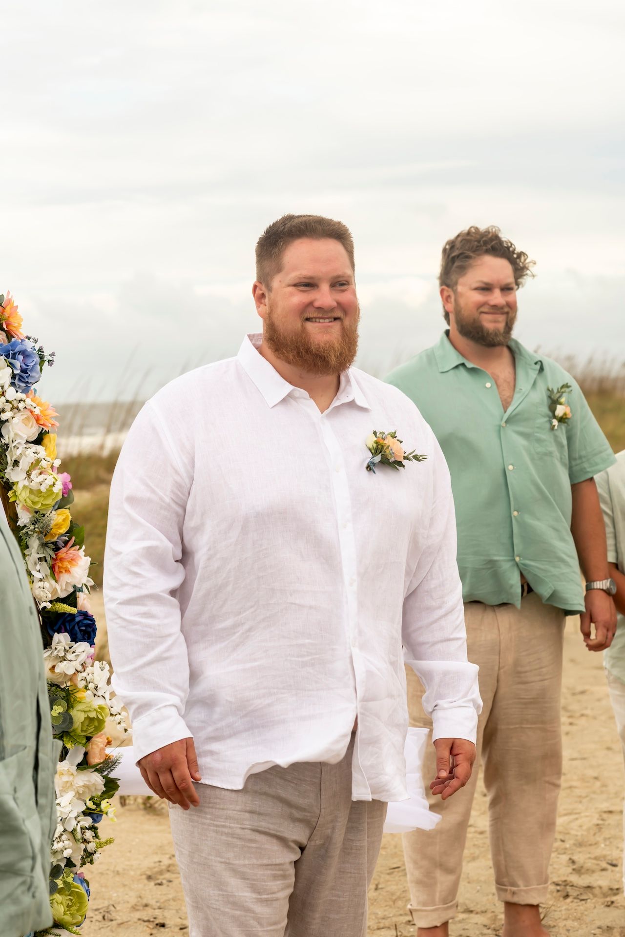 Two groomsmen wearing casual beach wedding attire stand on a sandy beach with floral decorations visible.
