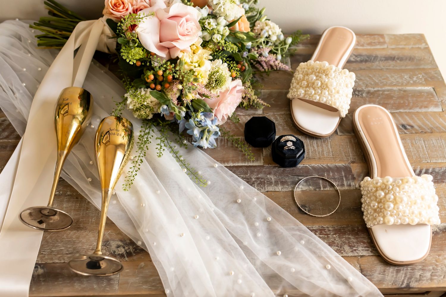 Wedding accessories including champagne flutes, bridal sandals, veil and pastel pink floral bouquet on rustic wooden surface.