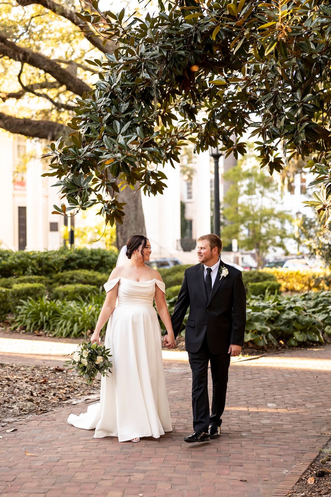 A wedding couple strolls hand in hand on a red brick path beneath a magnolia tree in an outdoor garden setting.