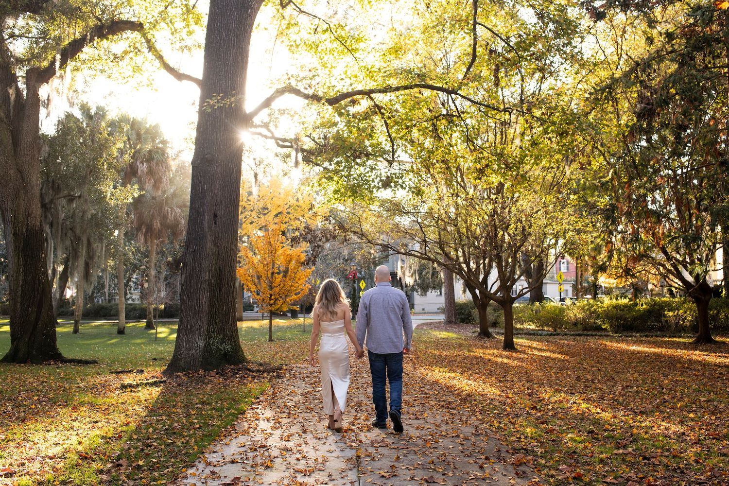 Two people holding hands while walking down a leaf-covered path in a sunlit autumn park.