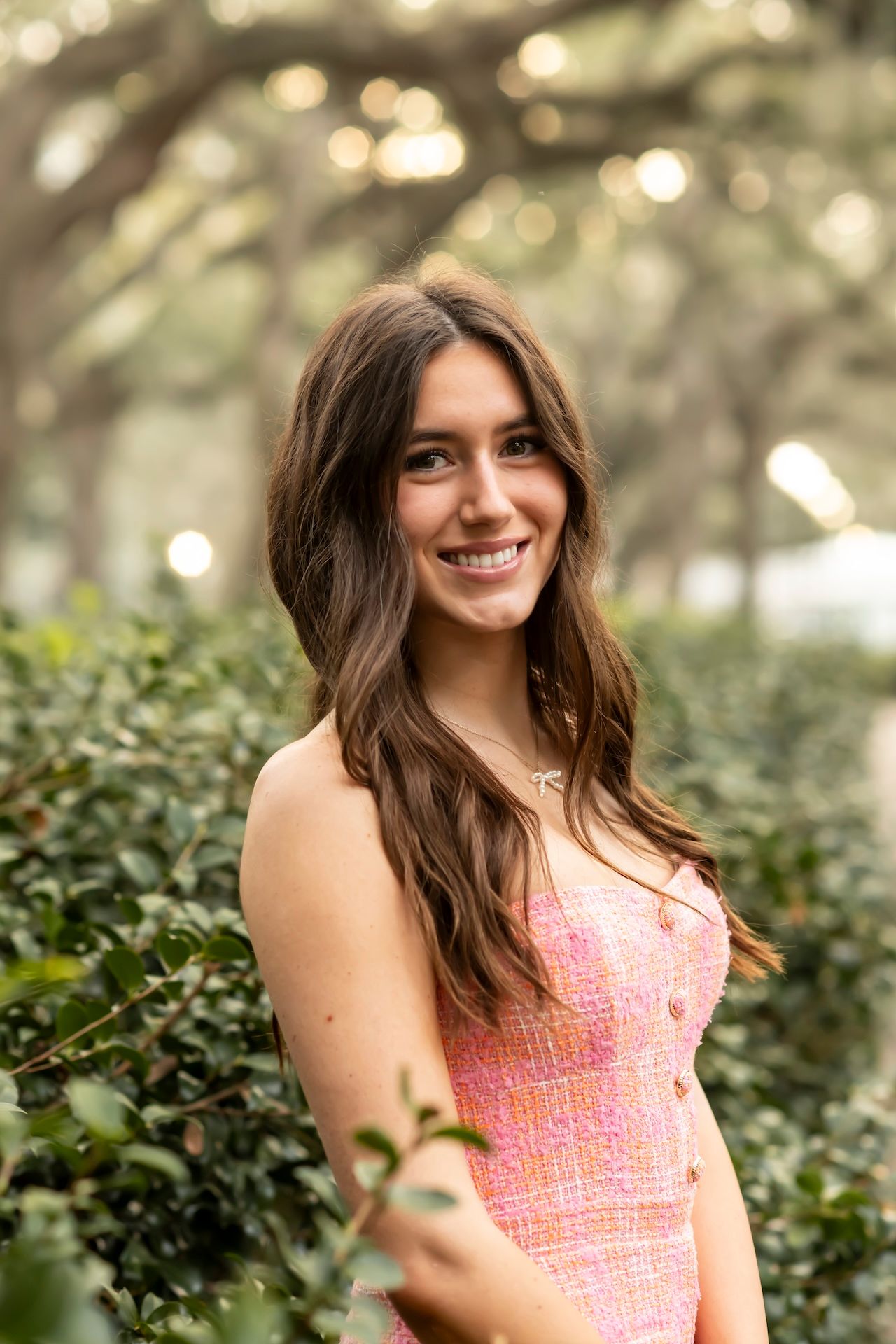 Portrait of someone in a pink patterned dress standing among green foliage with soft bokeh lights in the background.