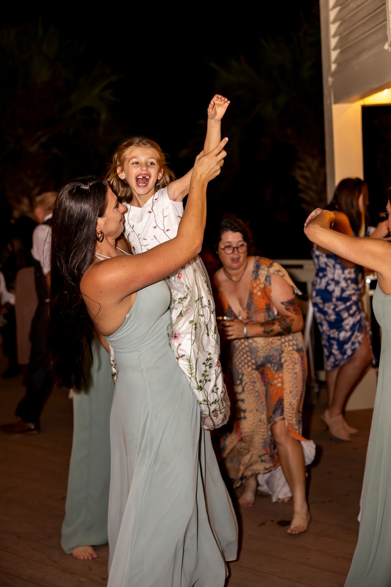 Joyful wedding guests dancing together at a nighttime reception on a covered patio.